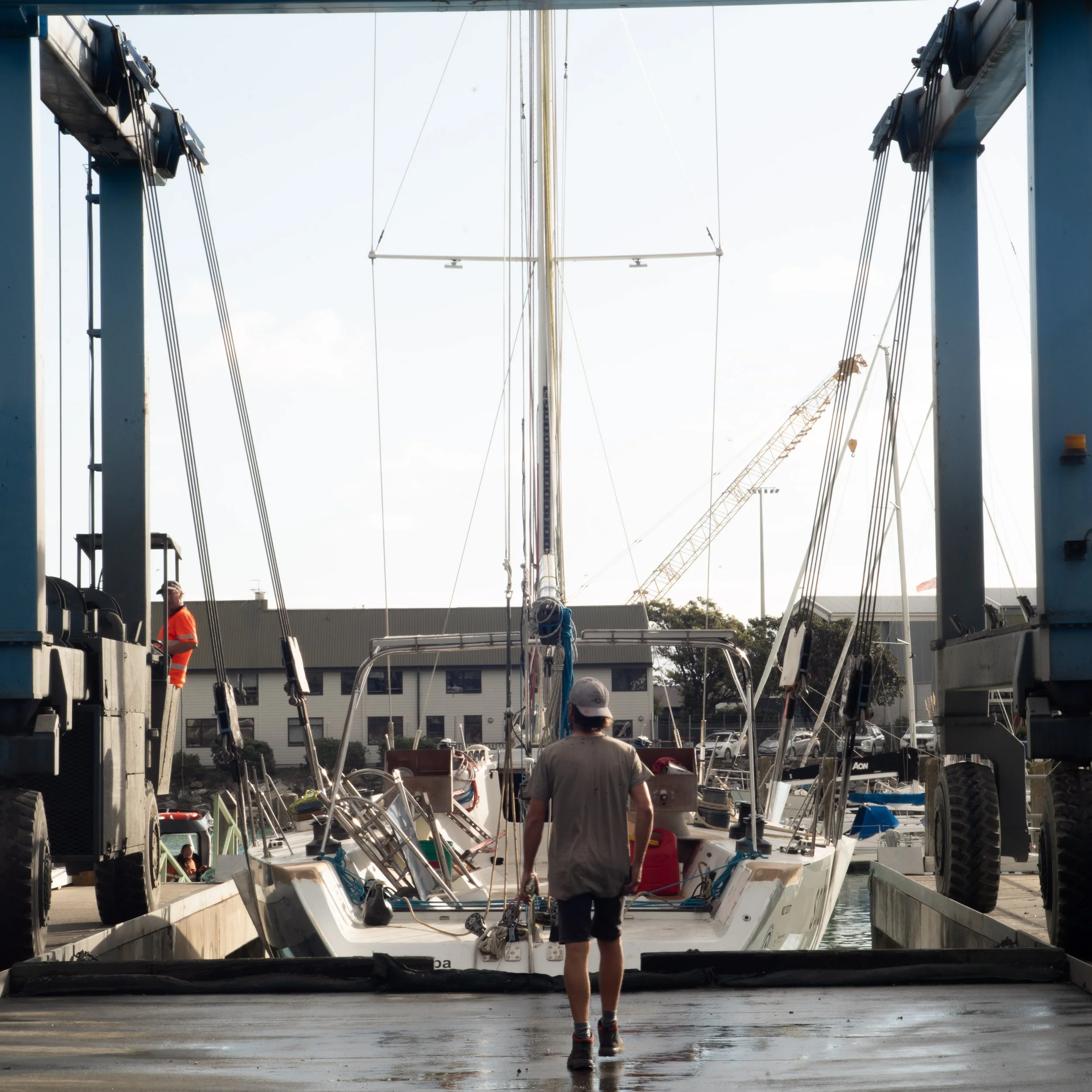 Person walking towards a docked sailboat at marina with other boats and buildings in background.