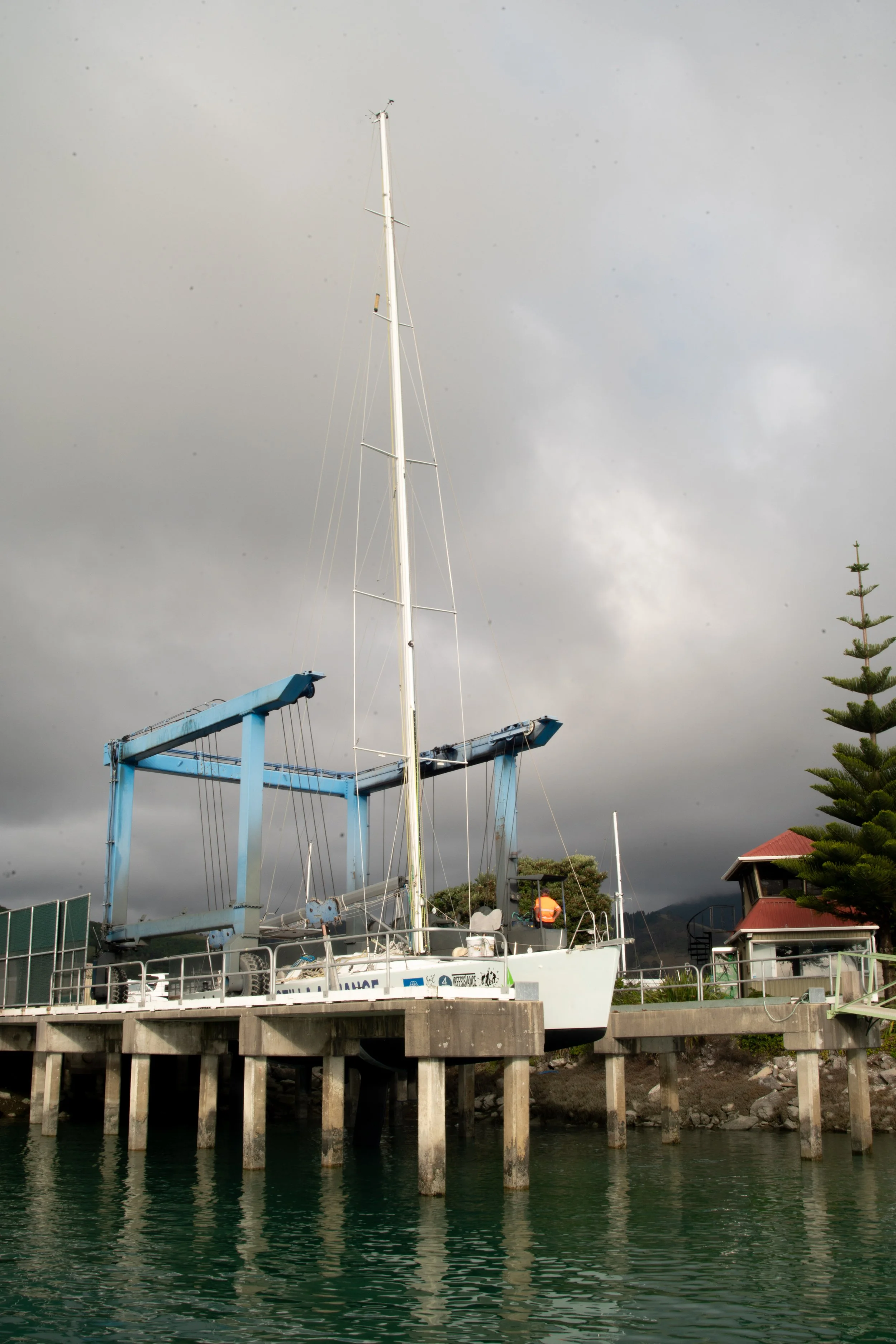 A sailboat docked at a pier with a crane and a small building nearby, under a cloudy sky.