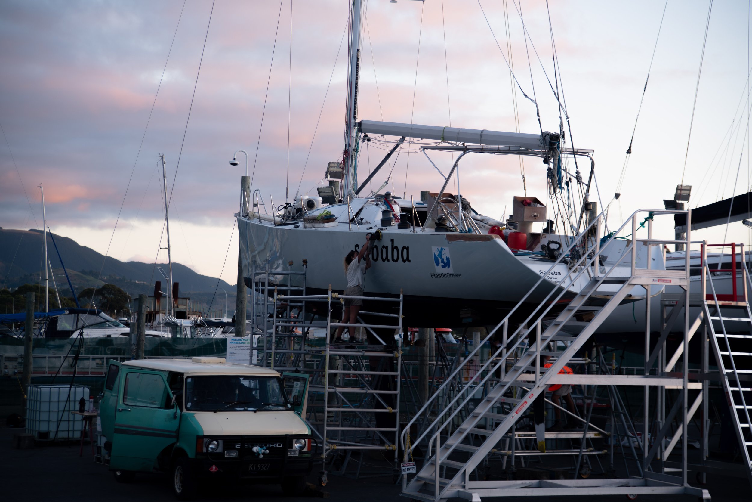 A sailboat named 'Sohaba' is on a dry dock surrounded by scaffolding, with a person working on it. There are other boats and a moored boat with a mountainous background at sunset.