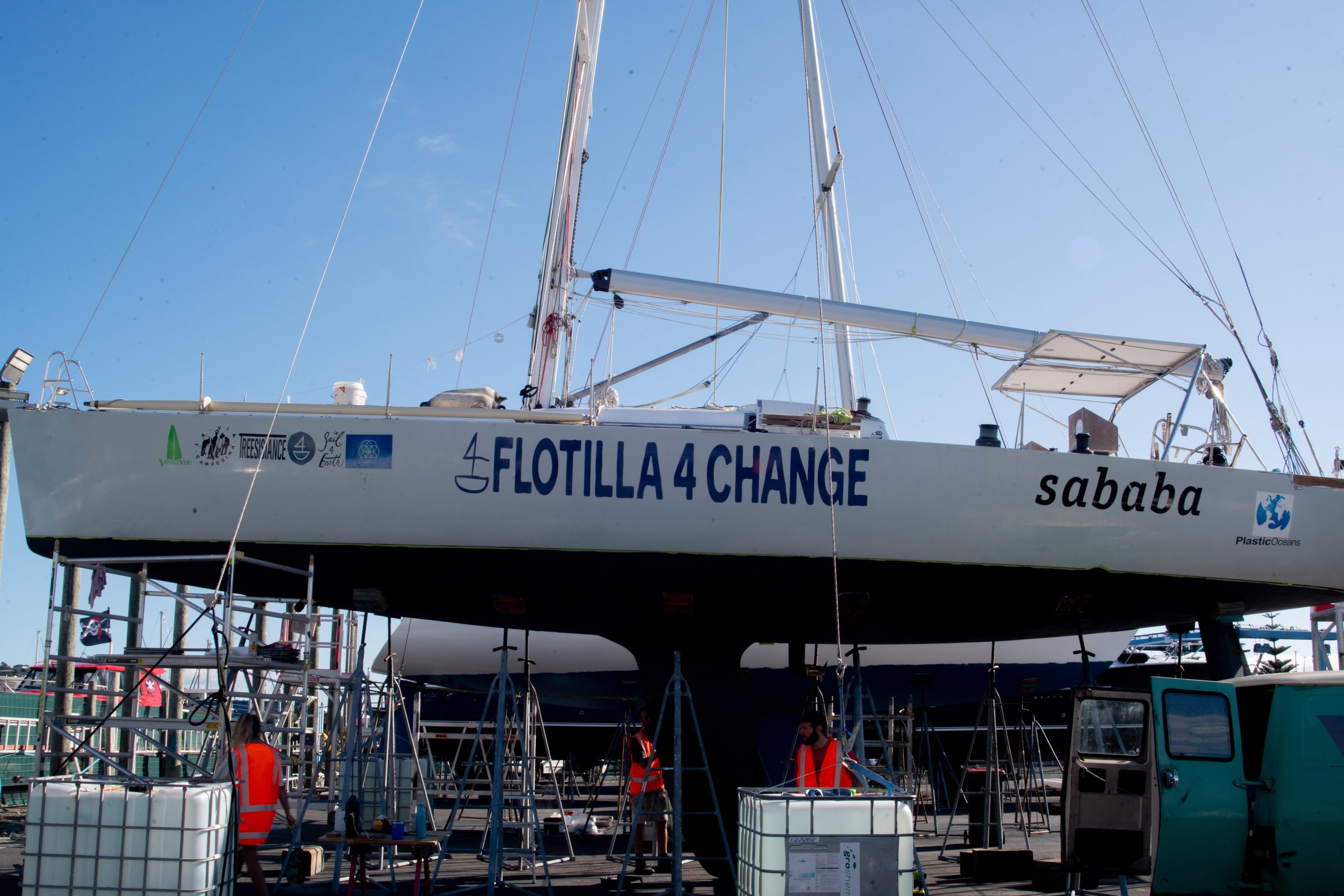 A sailboat named Flotilla 4 Change is on land, supported by scaffolding and surrounded by people in safety vests. The boat has various stickers and logos, and equipment is visible around it under a clear blue sky.