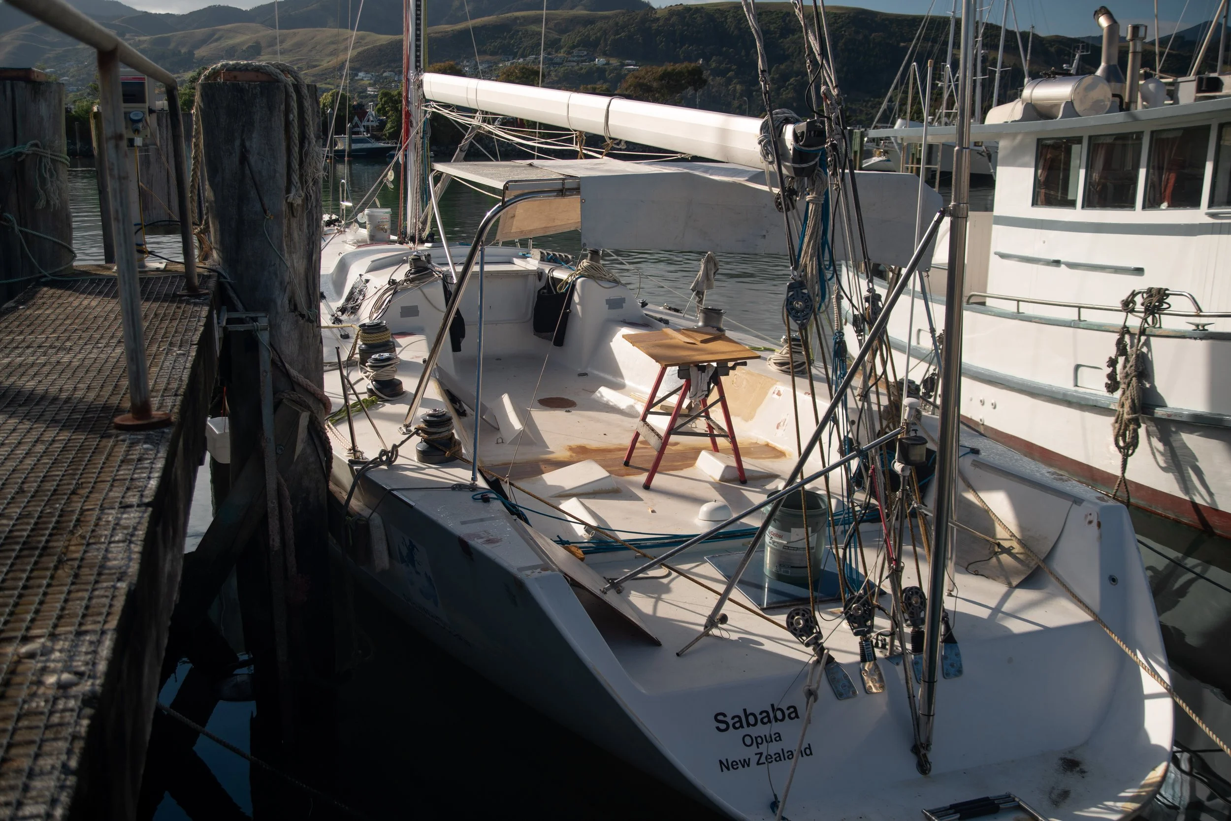 A sailboat docked at a marina with visible ropes, pulleys, and a small table on the deck. The boat has the name 'Sababa' and is registered in Opua, New Zealand, with mountains in the background.