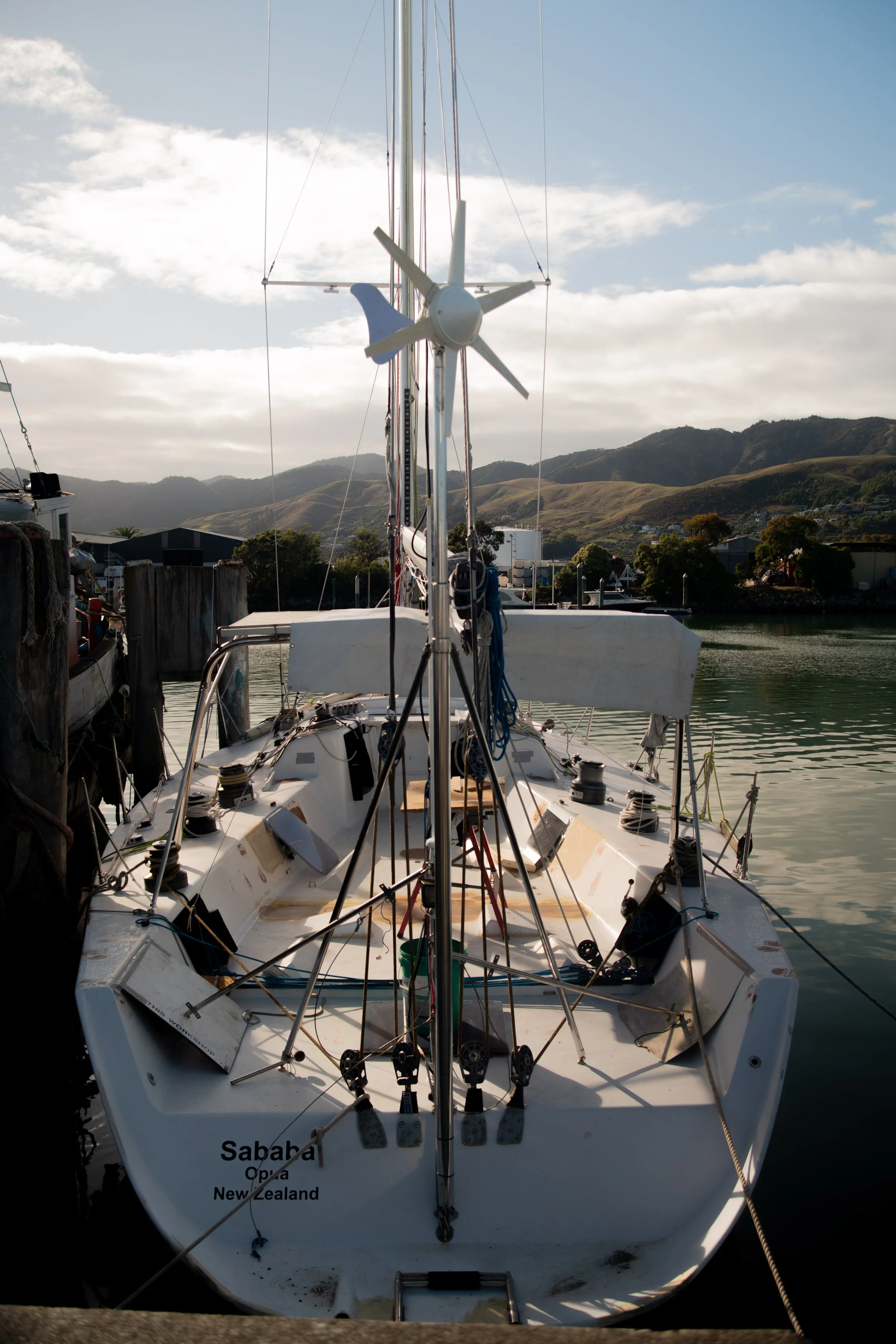 A sailboat docked at a marina, showing the bow with the name 'Sabaha' and 'Opua New Zealand', with surrounding mountains and partly cloudy sky in the background.