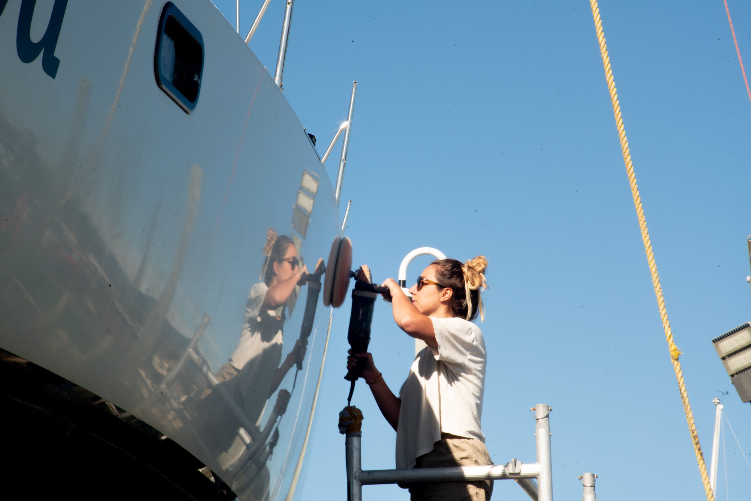 A woman with sunglasses and a bun hairstyle is polishing the side of a boat with a power buffer. She is standing on a small metal platform against a clear blue sky, with boat masts and ropes visible in the background.