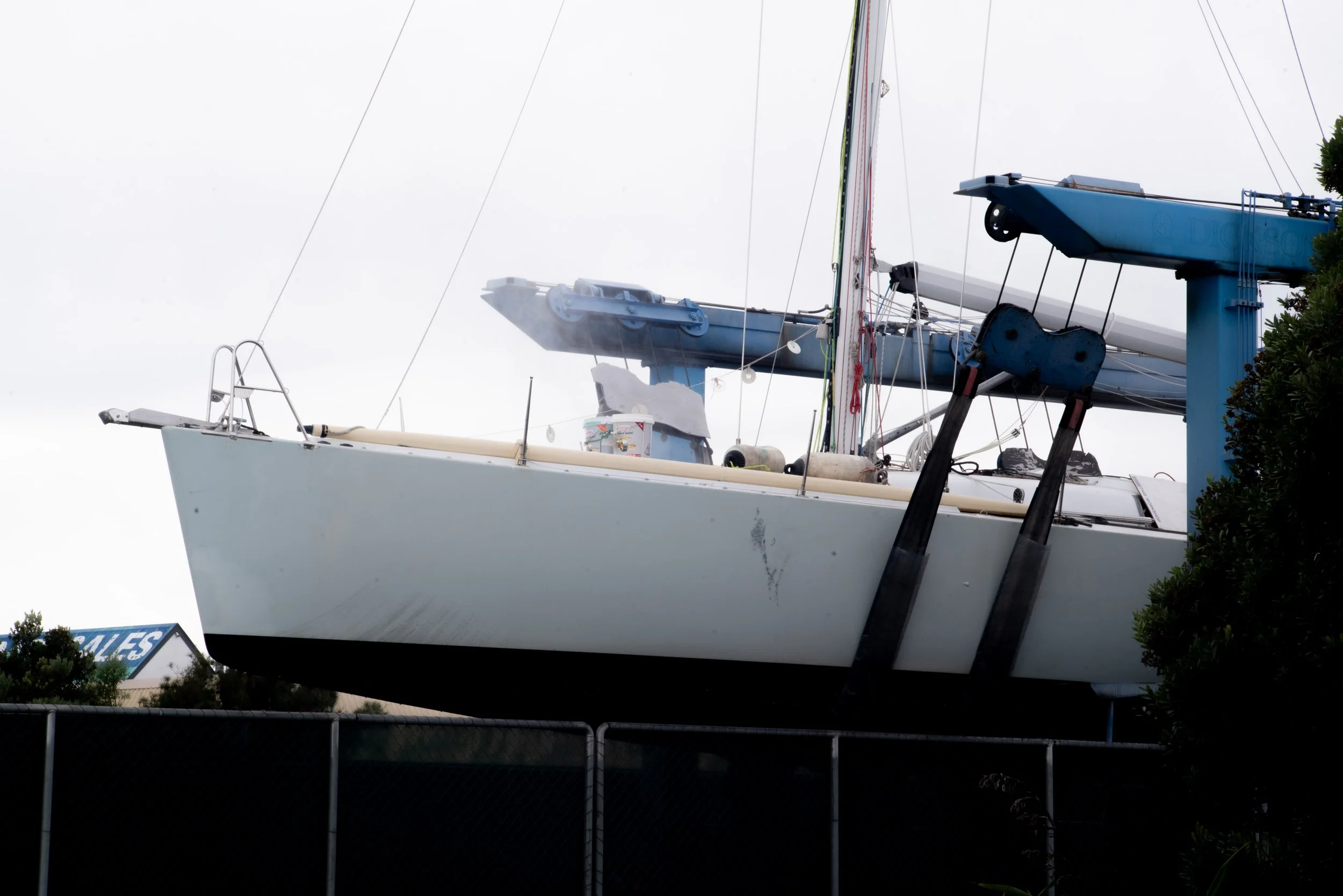 A sailboat on dry dock with a blue crane lifting it, overcast sky.