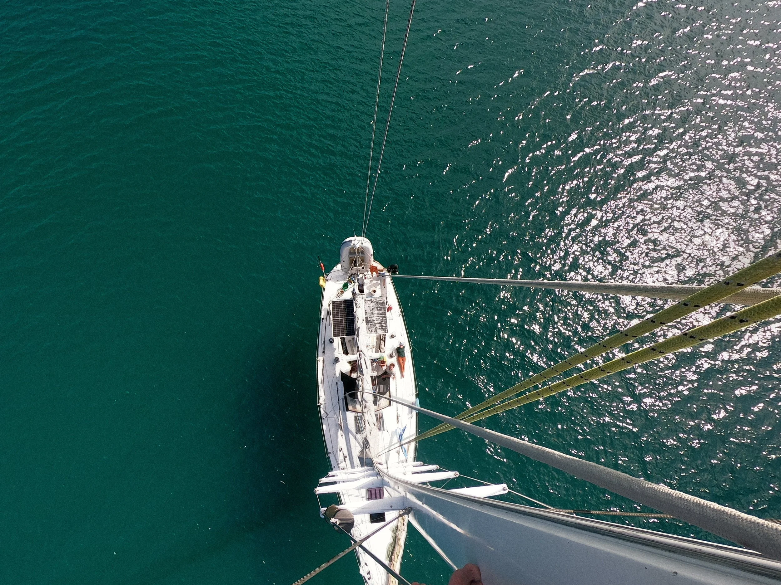 Overhead view of a sailboat on turquoise water with sunlight reflecting off the surface, taken from the top of the mast.