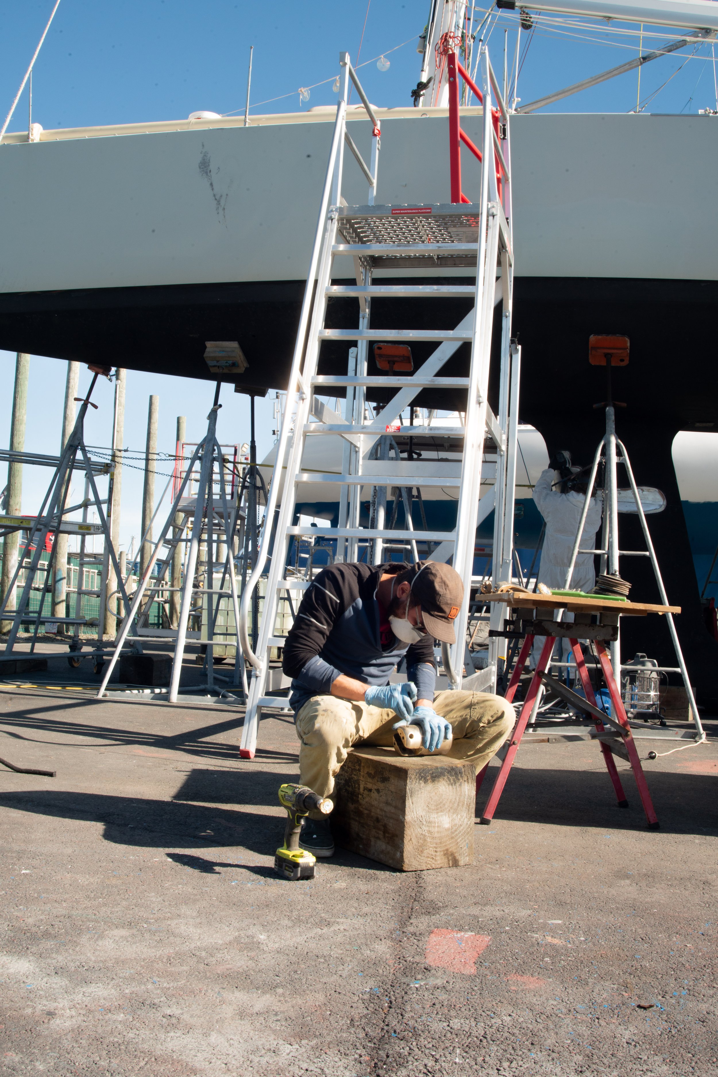 A person working on a wooden block under a large boat at a boatyard, with scaffolding and tools surrounding them.