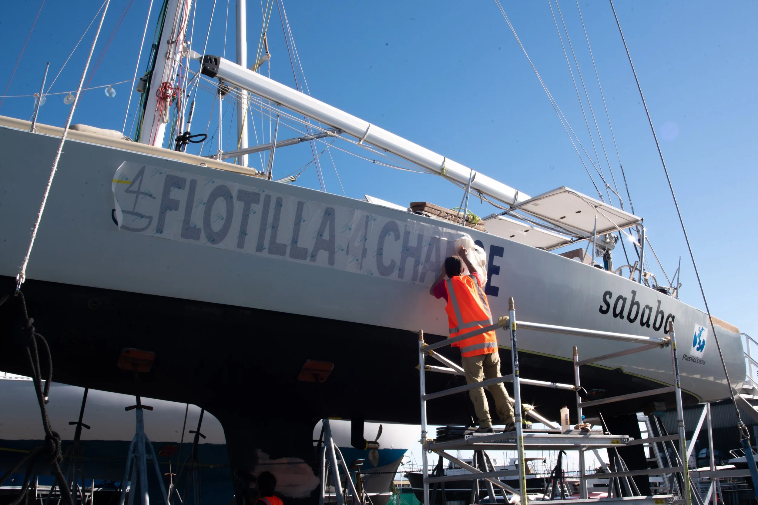 A person in an orange safety vest cleaning or painting the side of a large white sailboat named 'sababa' at a boatyard, with a clear blue sky in the background.