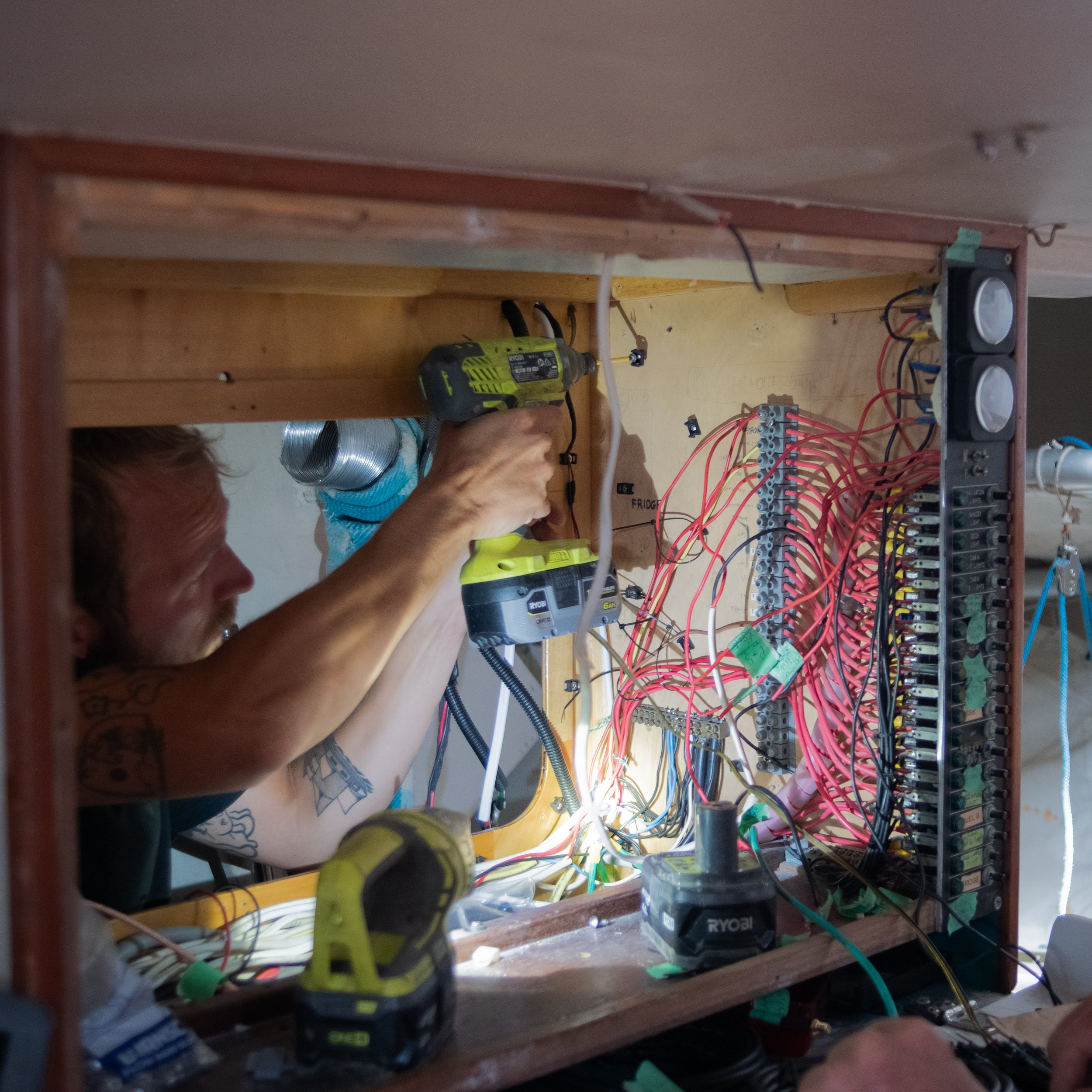Person working on electrical wiring inside a wooden panel box, using a power drill, with numerous wires and circuit components visible.