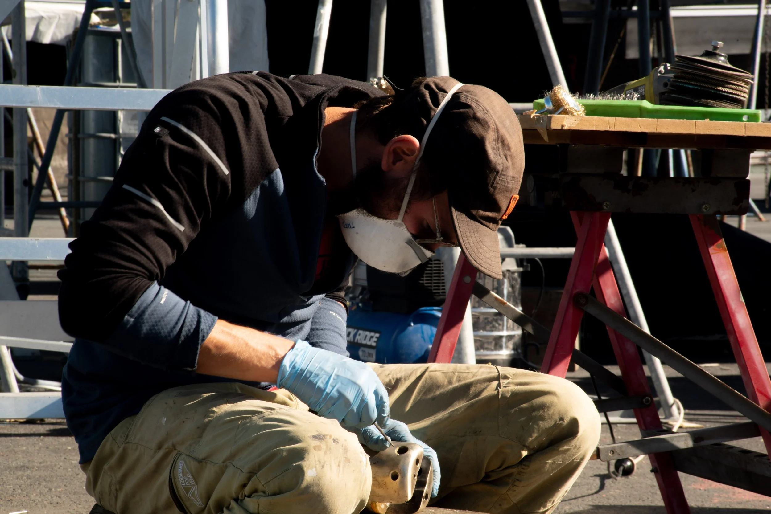 Man wearing a brown cap, glasses, face mask, black jacket, and beige pants working on a mechanical part outdoors, sitting on the ground with tools and a workbench nearby.