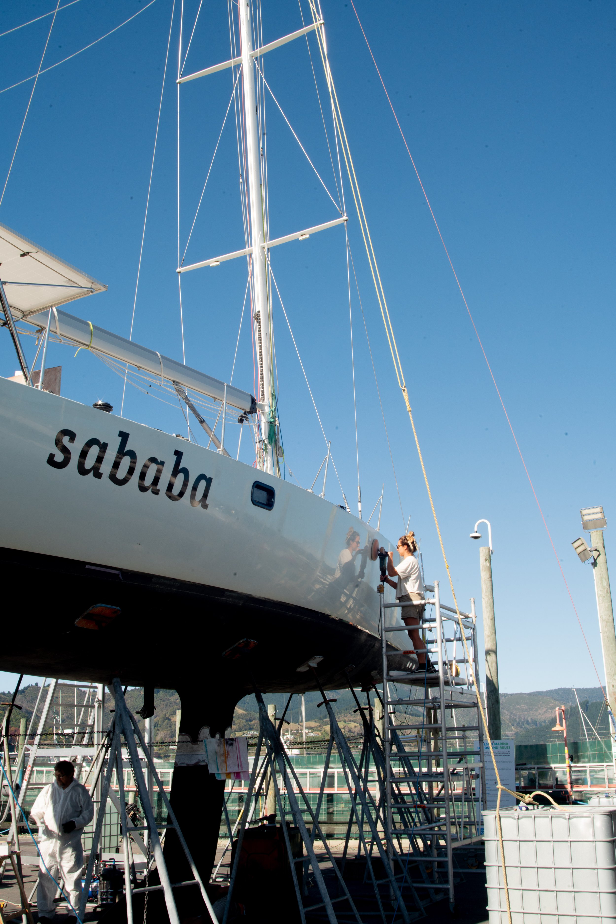 A woman polishing the hull of a sailboat named 'saba' at a marina during daytime with a clear blue sky.