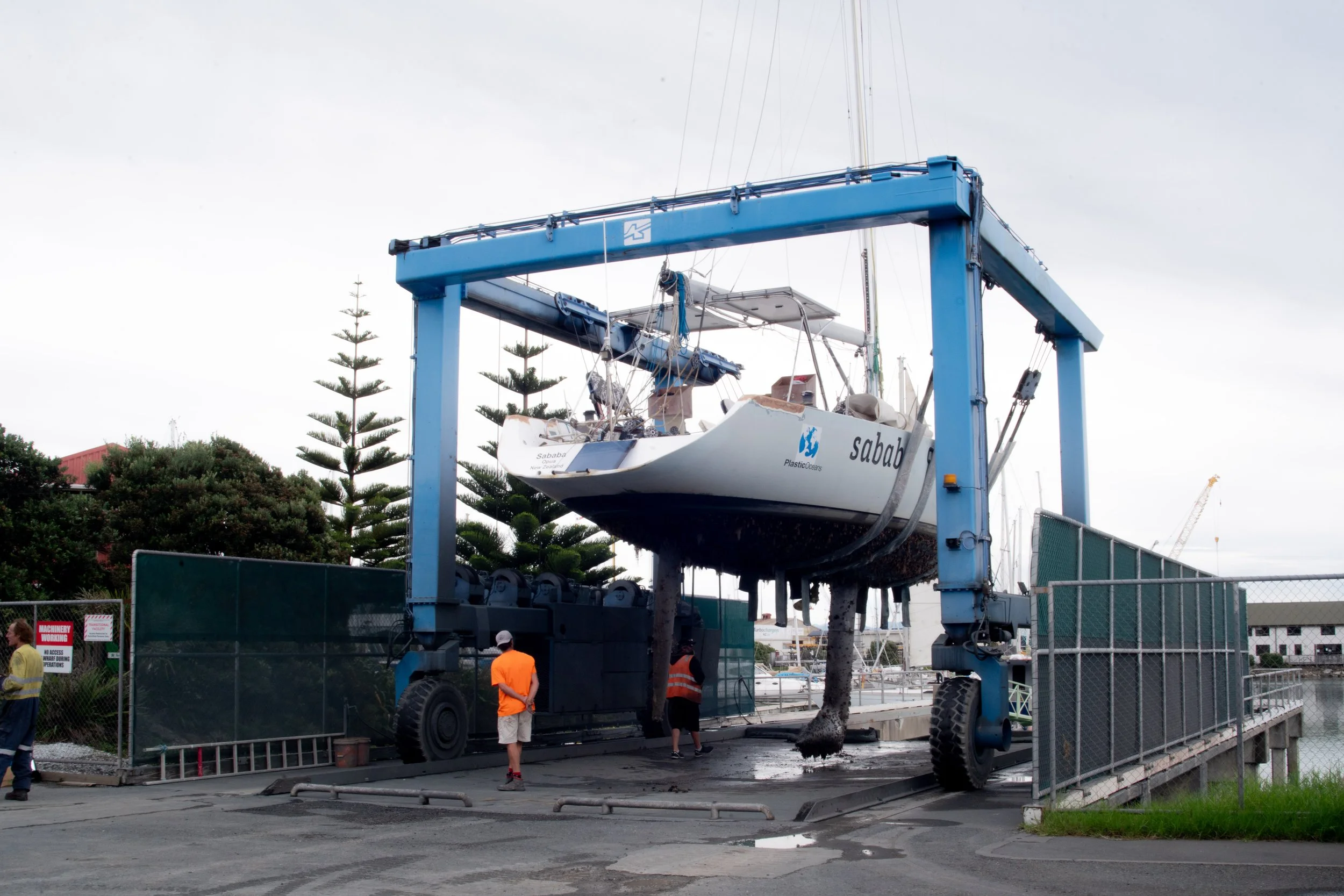 A boat being lifted out of the water by a large blue boat hoist at a marina. Several workers and onlookers are nearby.