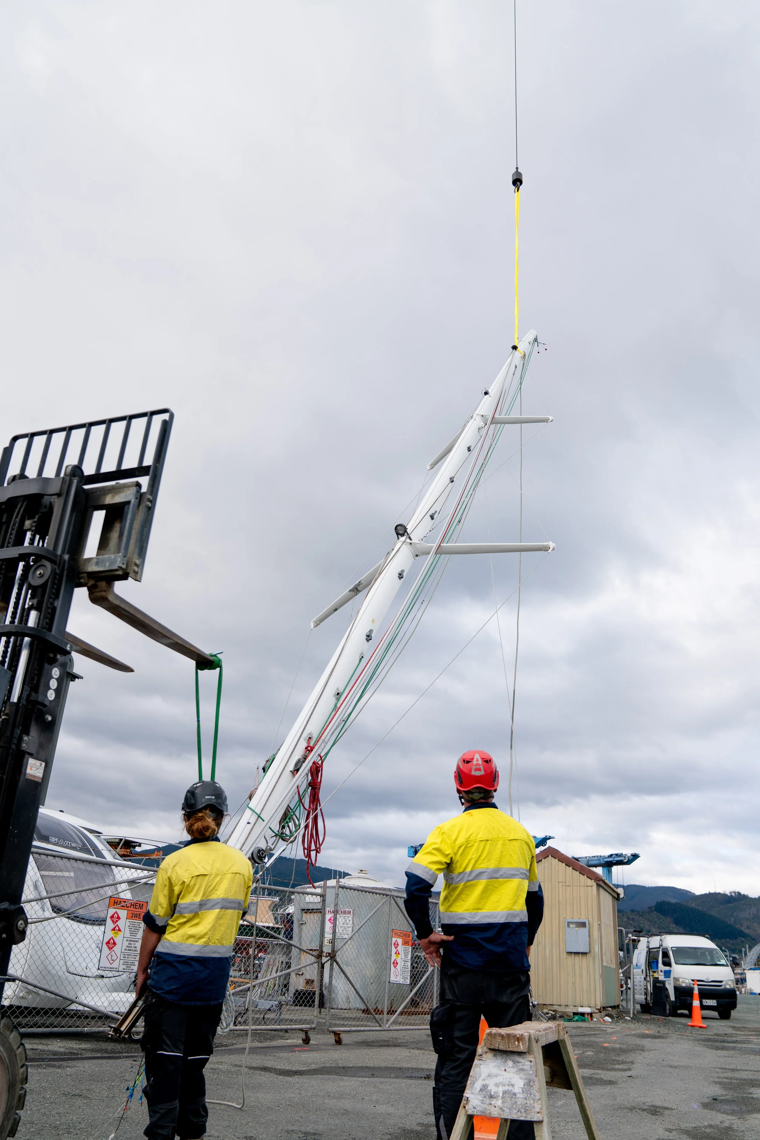 Two workers in yellow safety vests and helmets overseeing the lifting of a sailboat mast using a crane at a dock.