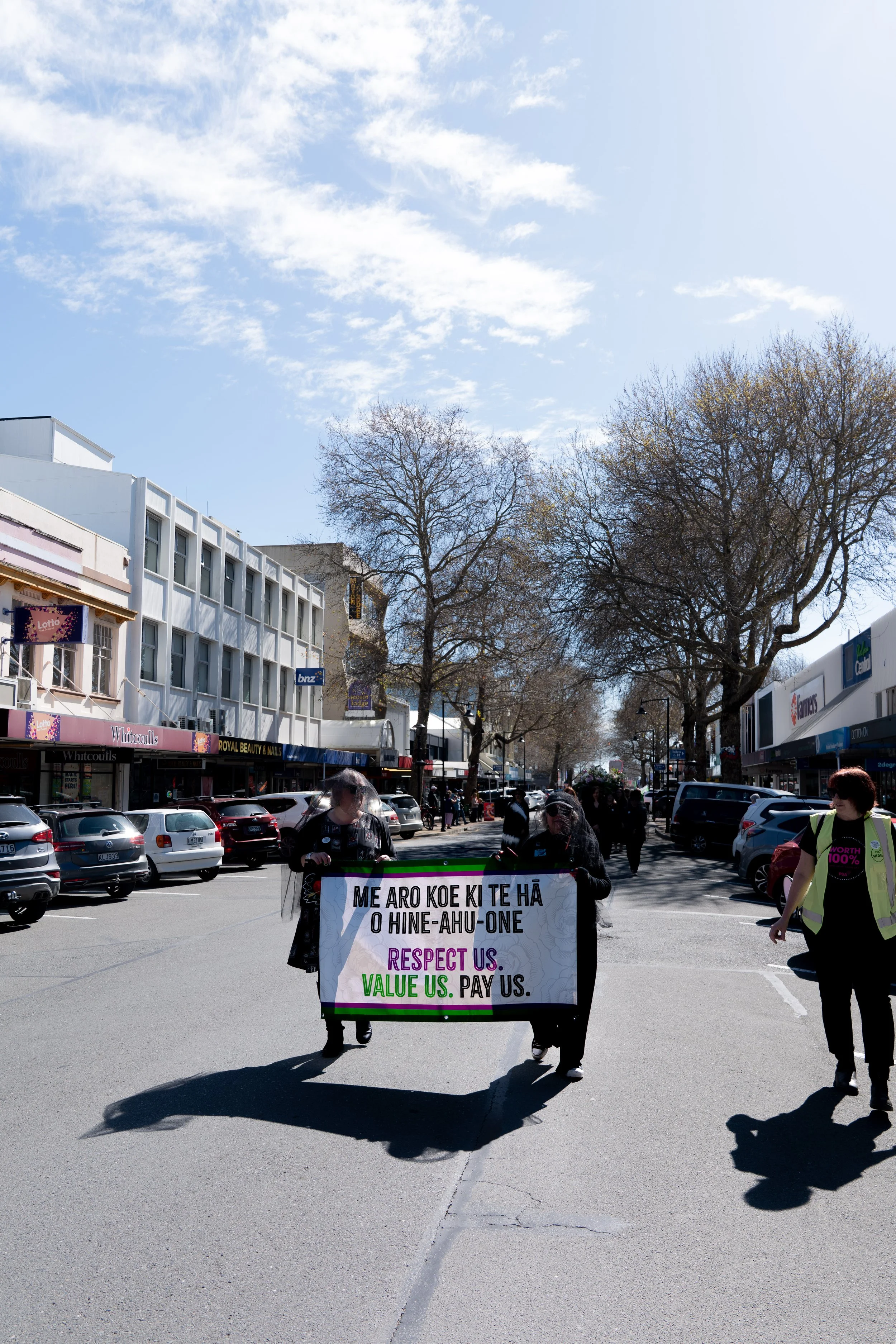 People marching on a city street holding a sign with a message in Maori language. The street is lined with parked cars and storefronts, with leafless trees and a partly cloudy sky overhead.
