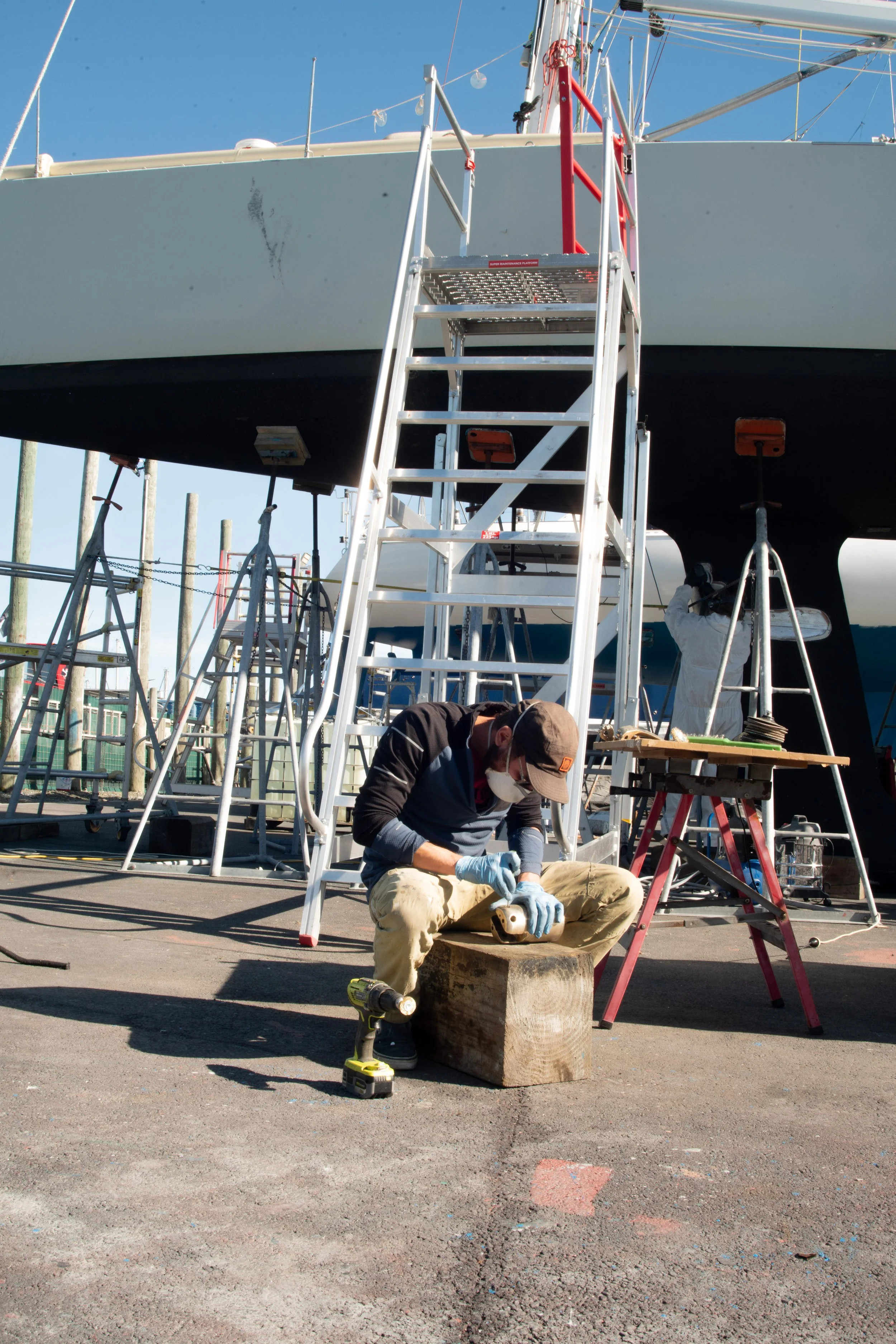 Man working on a boat at a marina, wearing gloves, a cap, and a face mask, with tools and equipment around him.