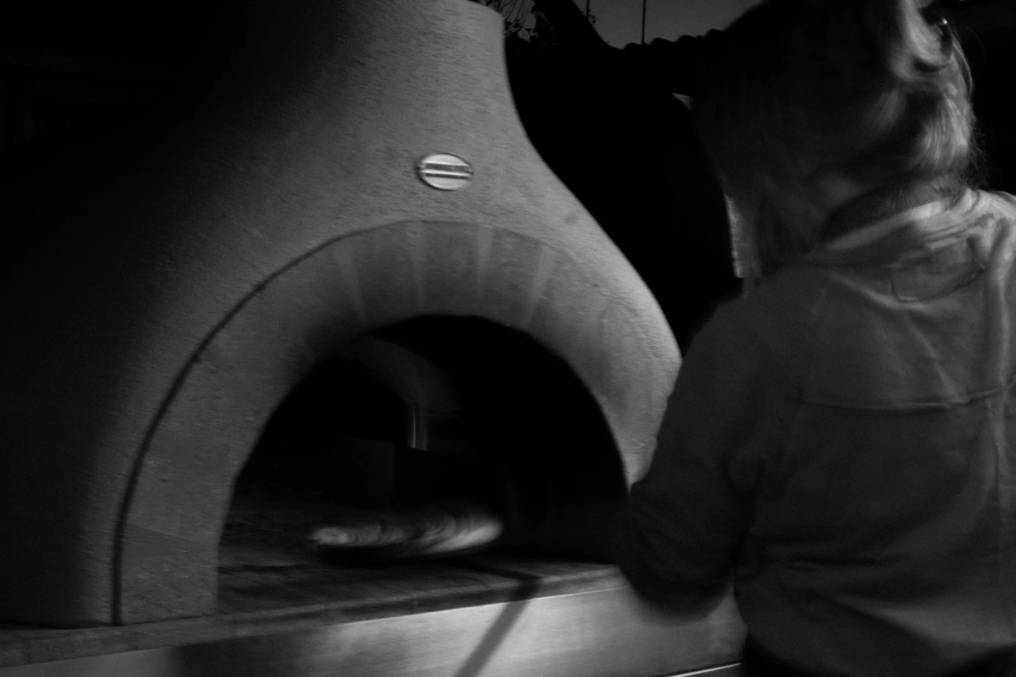 A person with long hair wearing a jacket is placing a pizza inside a wood-fired pizza oven.