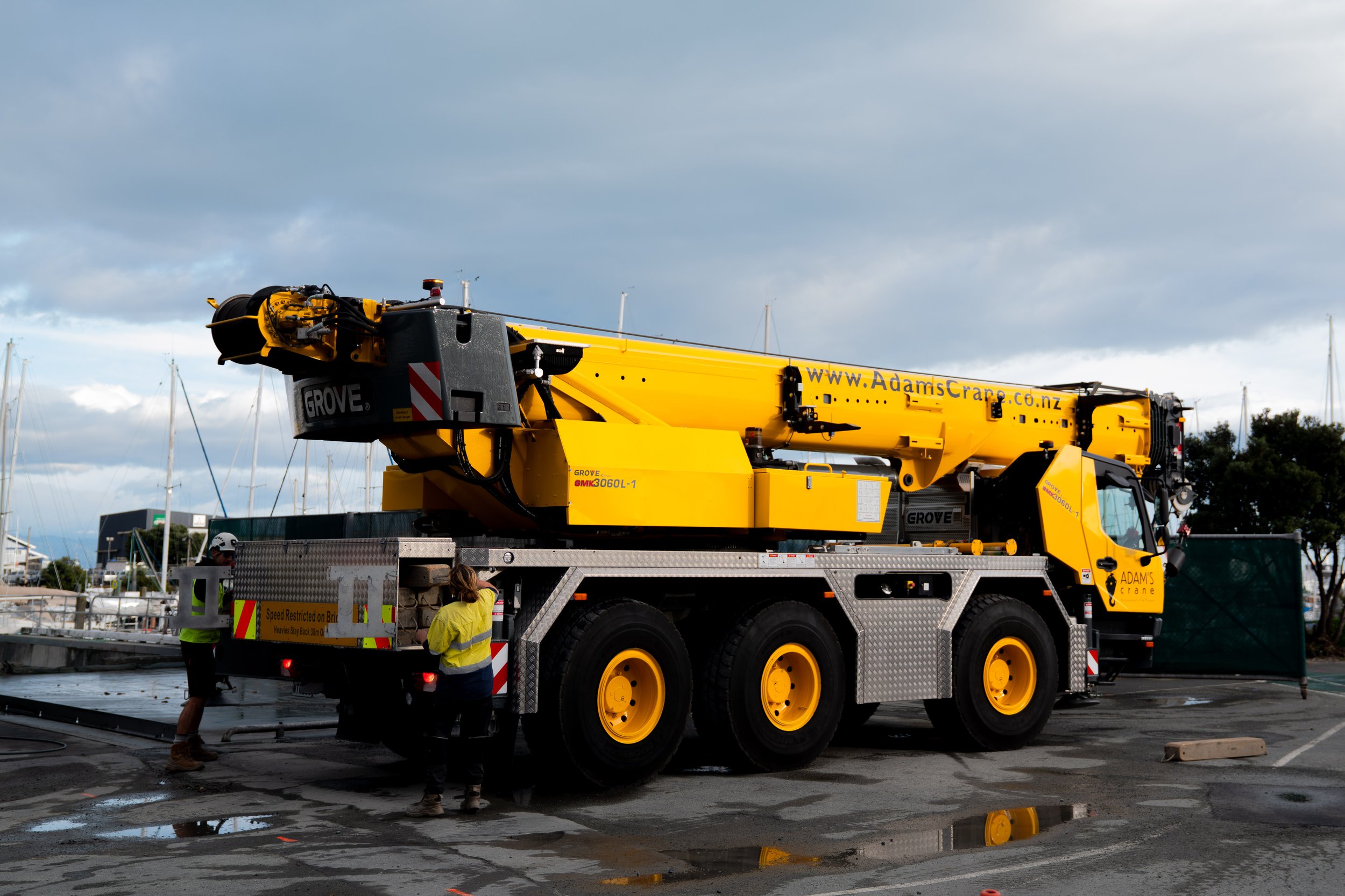 A large yellow crane truck with the branding 'Grove' and 'Adams Crane' on it. The vehicle is parked on a wet surface with some puddles, with three workers in safety vests nearby. The background shows sailboats and a partly cloudy sky.