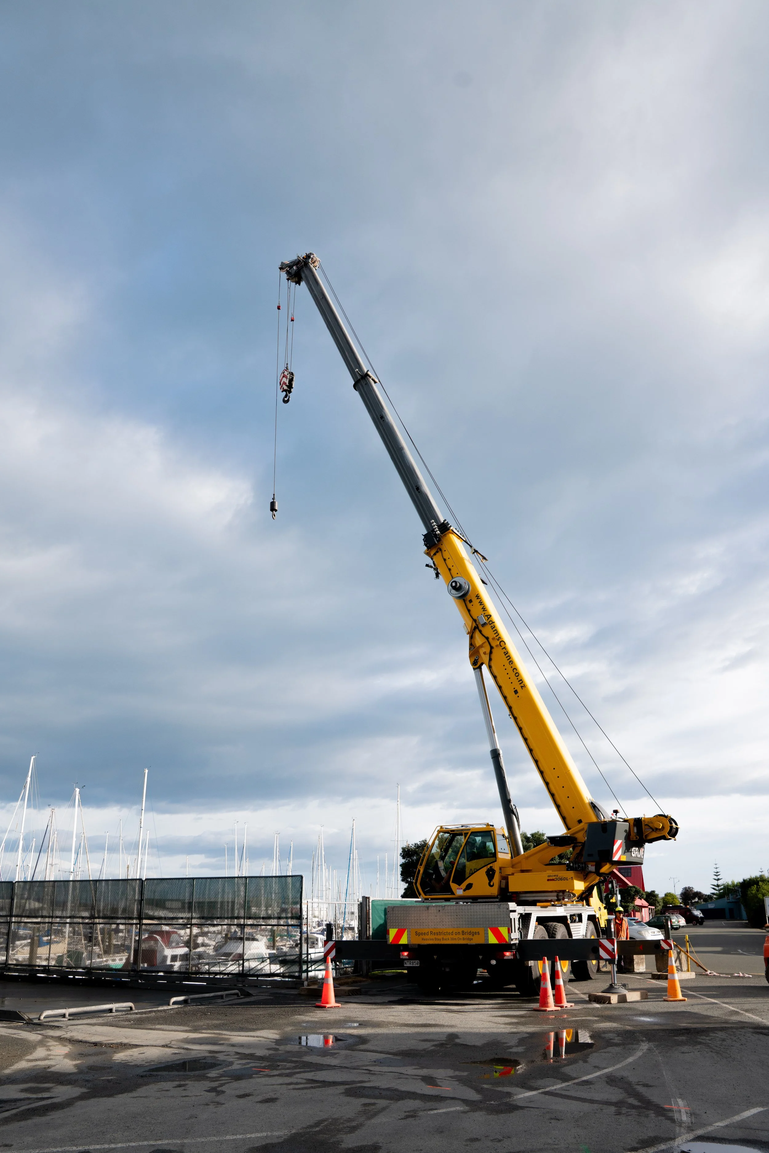 A large yellow crane lifting equipment at a construction site near a marina with sailboats.