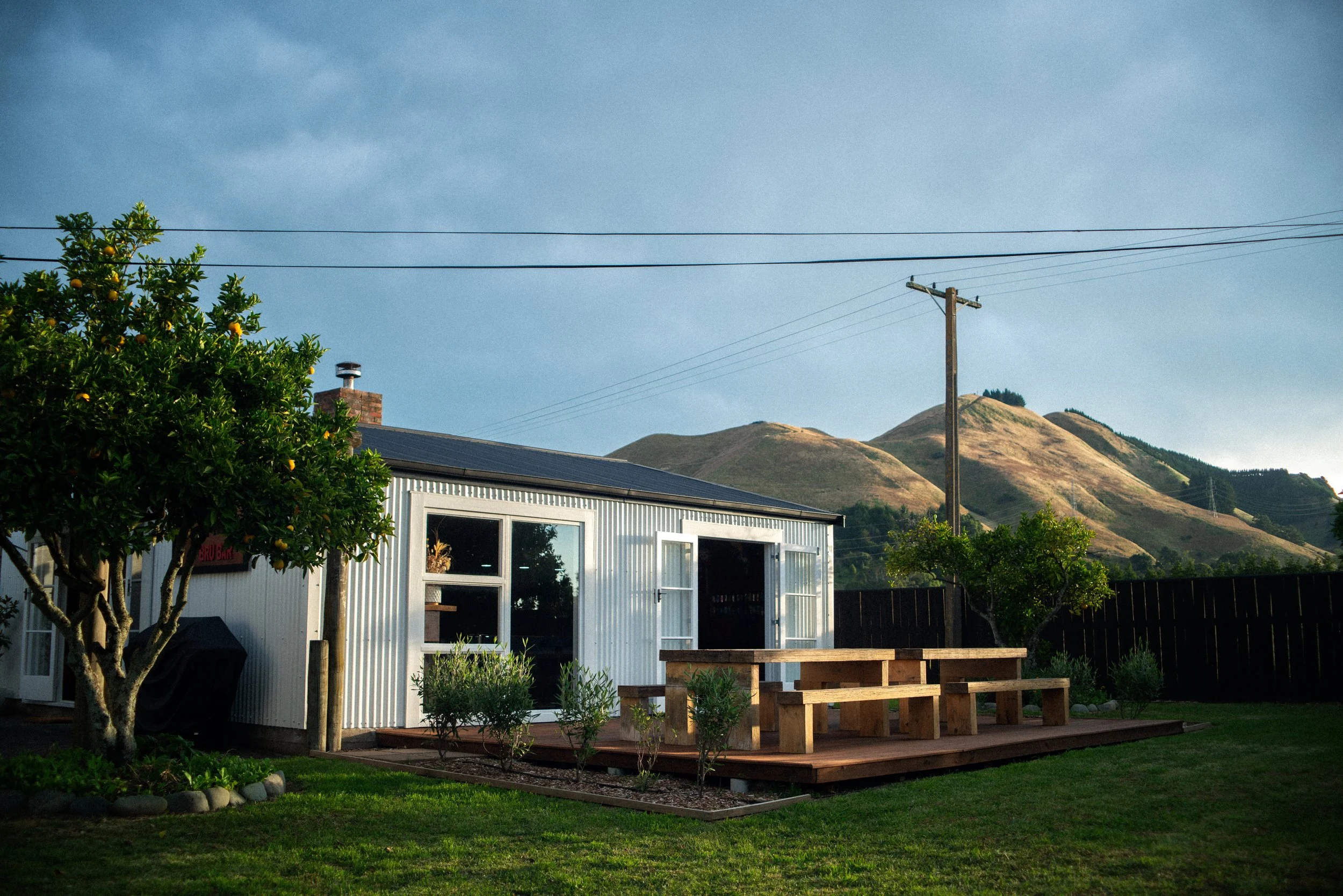 A small white building with large windows and an open door, situated in a lush green yard with trees and plants, against a backdrop of rolling hills and a blue sky.