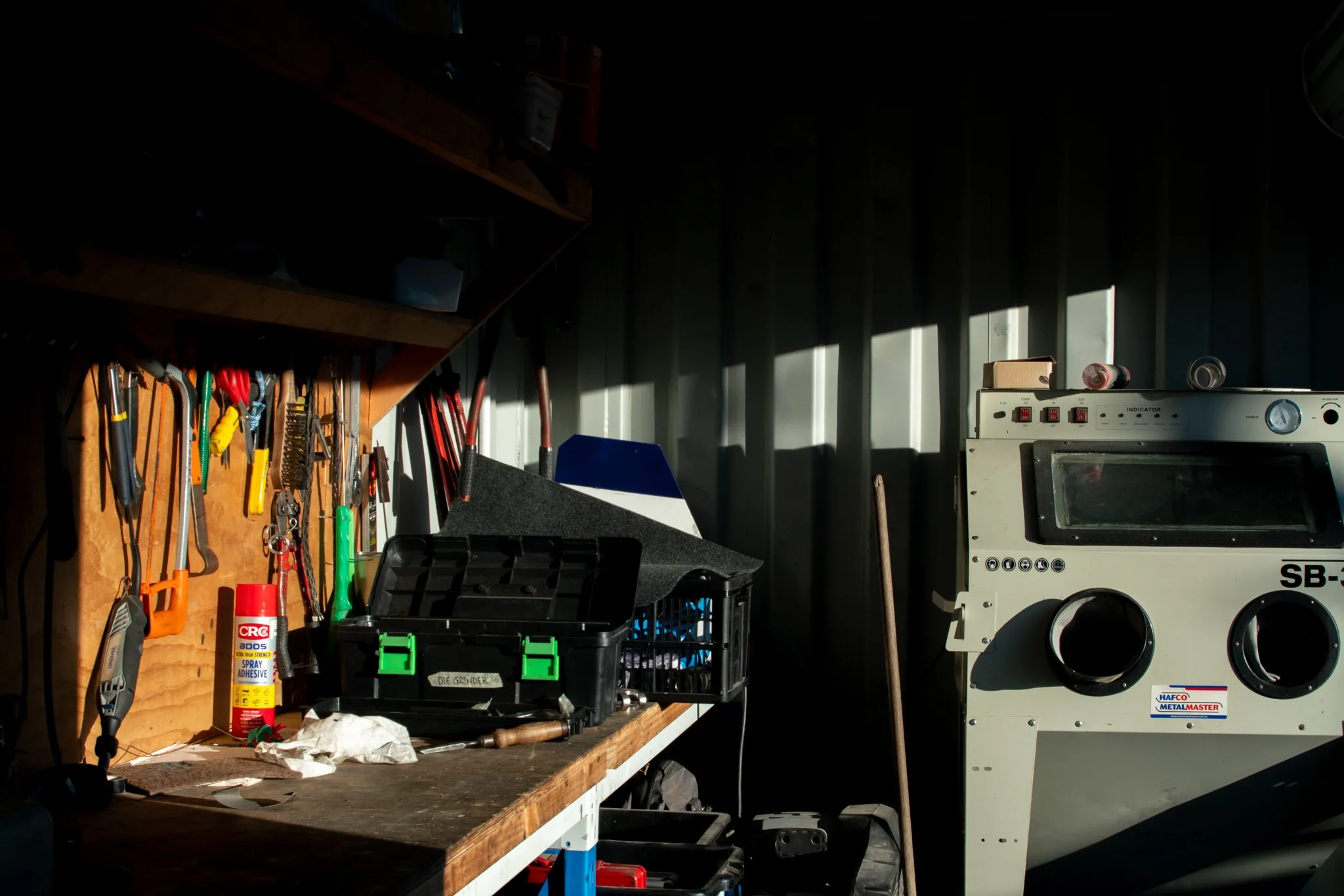 A cluttered workshop with a wooden workbench, various tools hanging on a pegboard, a spray adhesive can, and a large industrial machine against a corrugated metal wall.