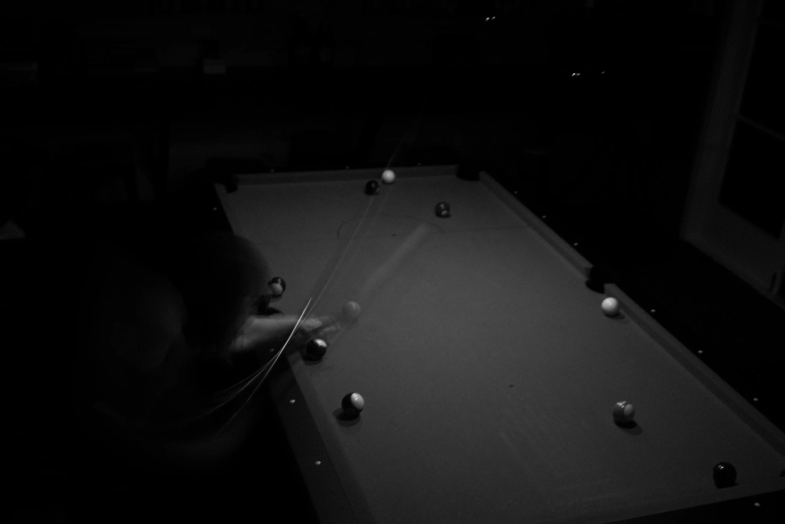A person playing pool in a dimly lit room, lining up a shot on a pool table with several balls scattered across the table.