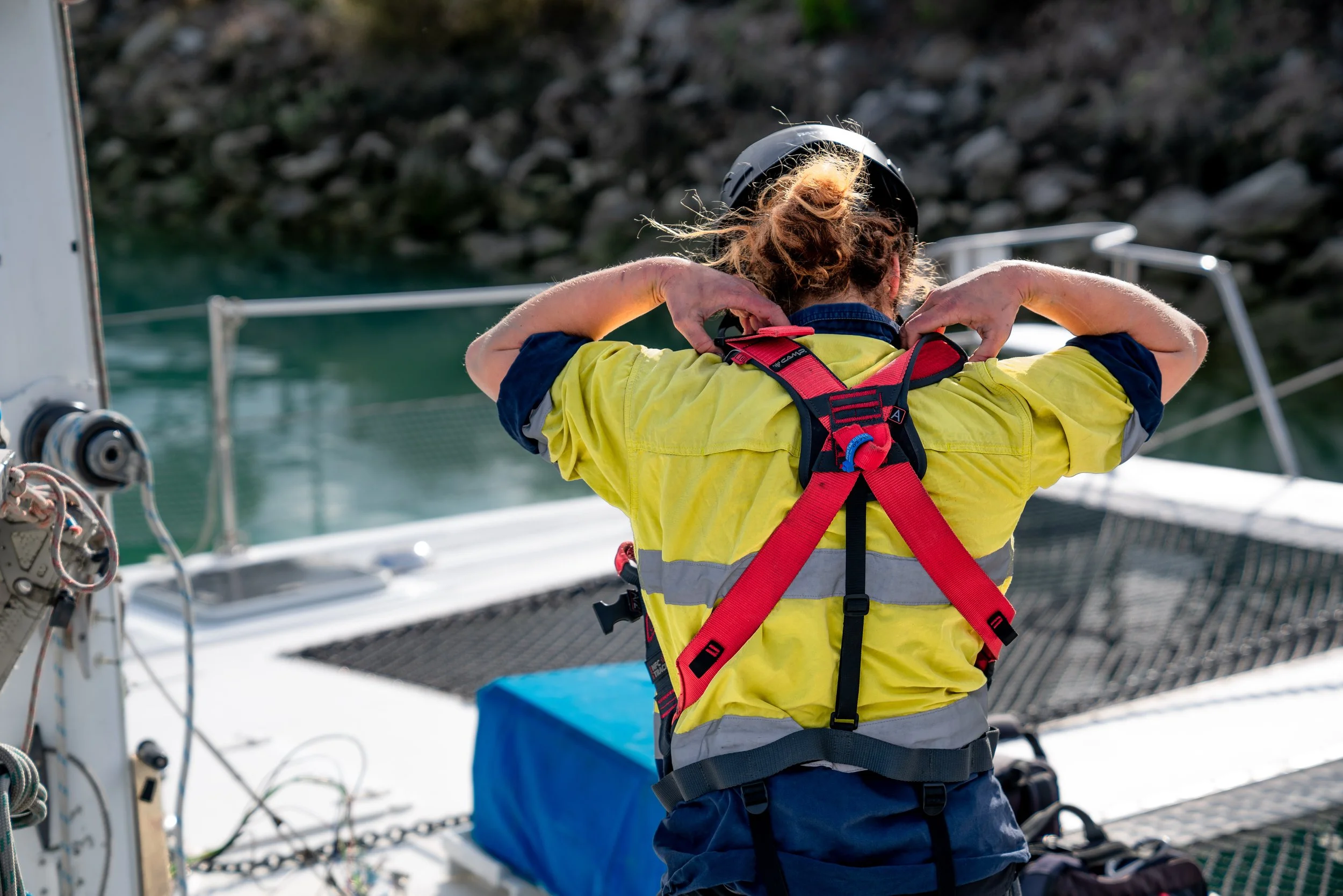A woman with curly hair wearing a yellow shirt, adjusting her life jacket on a boat docked near the water.