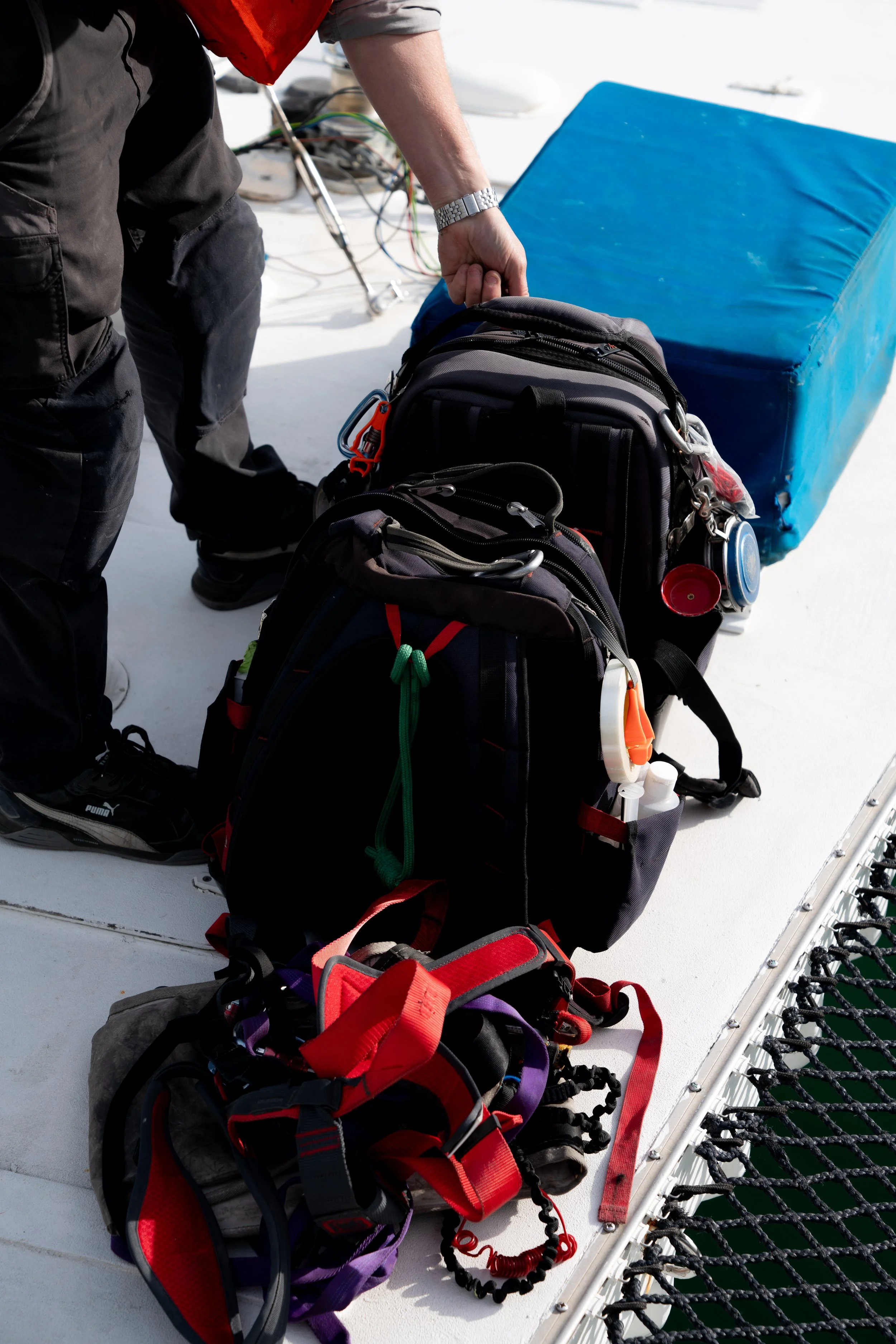 Person handling two backpacks and a harness on the deck of a boat, with a blue cushion and some wires in the background.