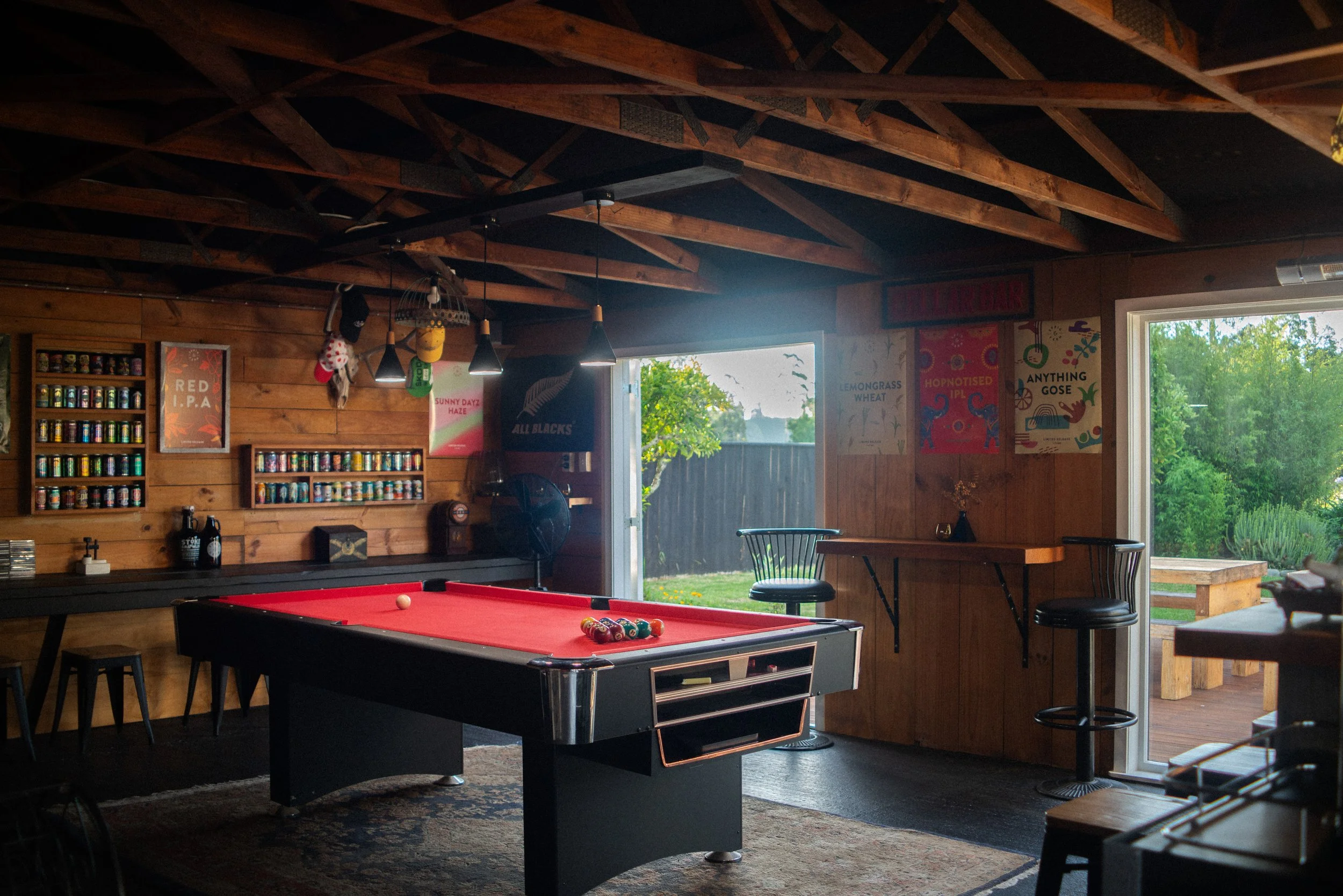 Interior of a cozy game room with a red pool table, wooden walls, and posters, illuminated by pendant lights; with large windows revealing outdoor greenery.