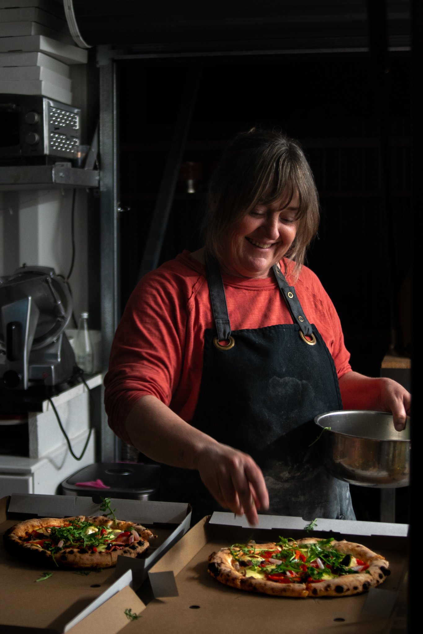 A woman in a red shirt and black apron preparing two pizzas with fresh greens on top in a kitchen.
