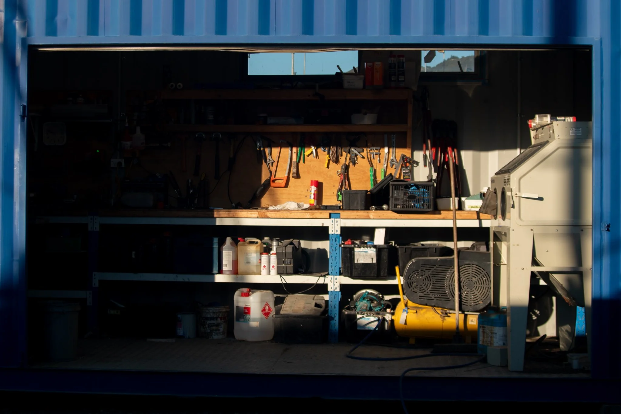 Inside a cluttered garage with tools hanging on a wooden board, shelves with various cans and containers, and a large industrial machine on the right side.