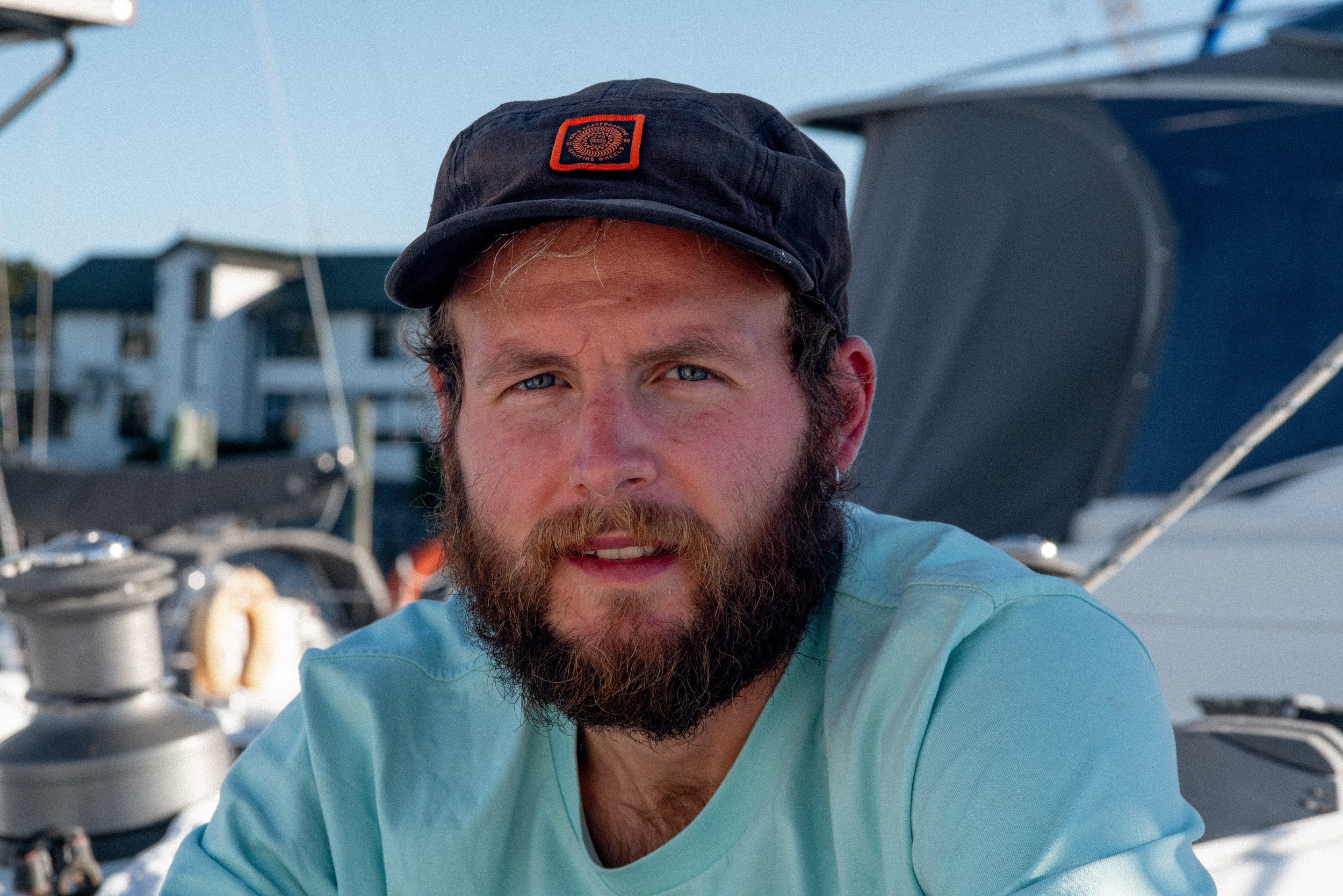 A man with a beard and blue eyes wearing a dark cap and light blue shirt, sitting on a boat with a marina in the background.