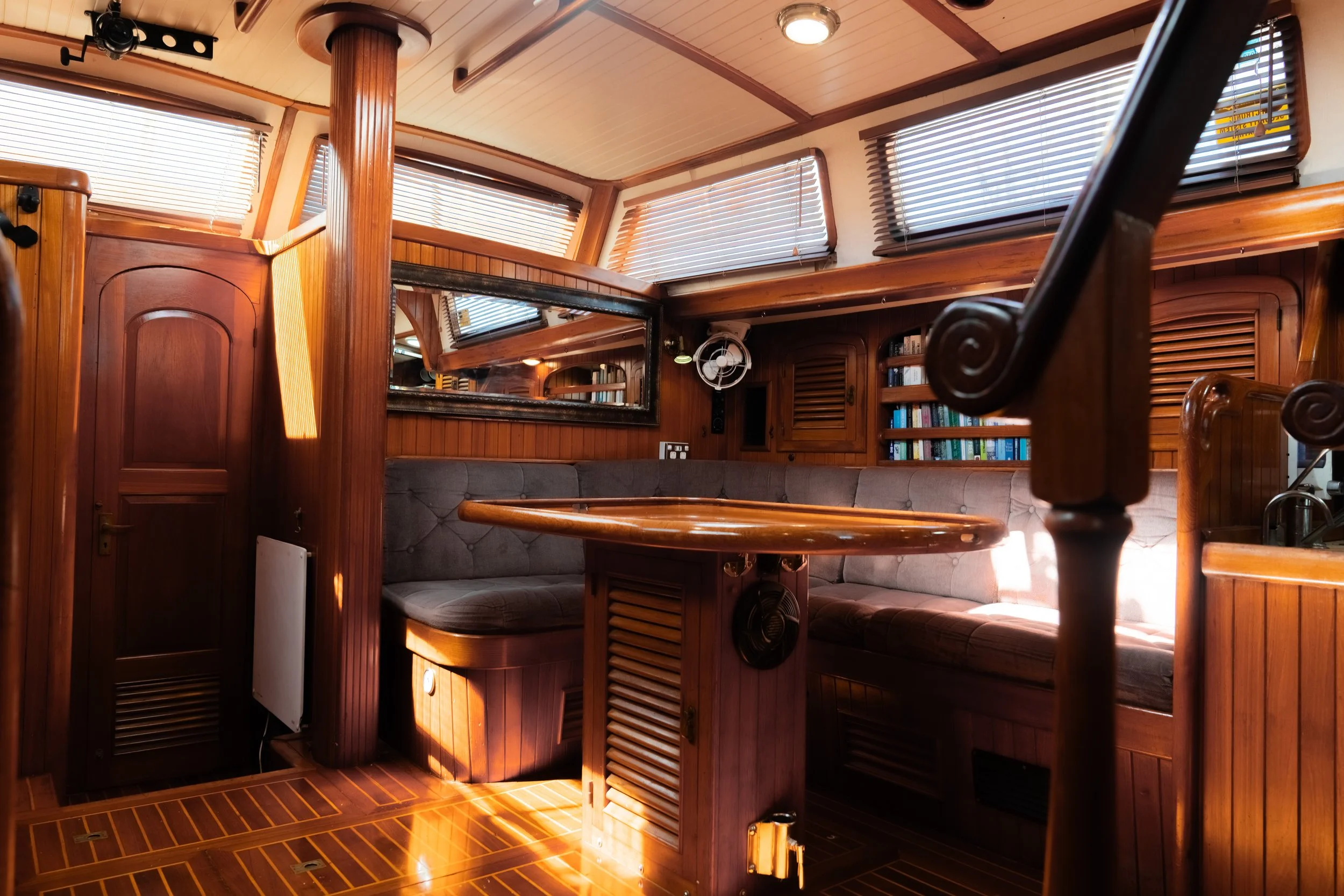 Interior of a wooden boat with a curved seating area, window blinds, a mirror, and a bookshelf.