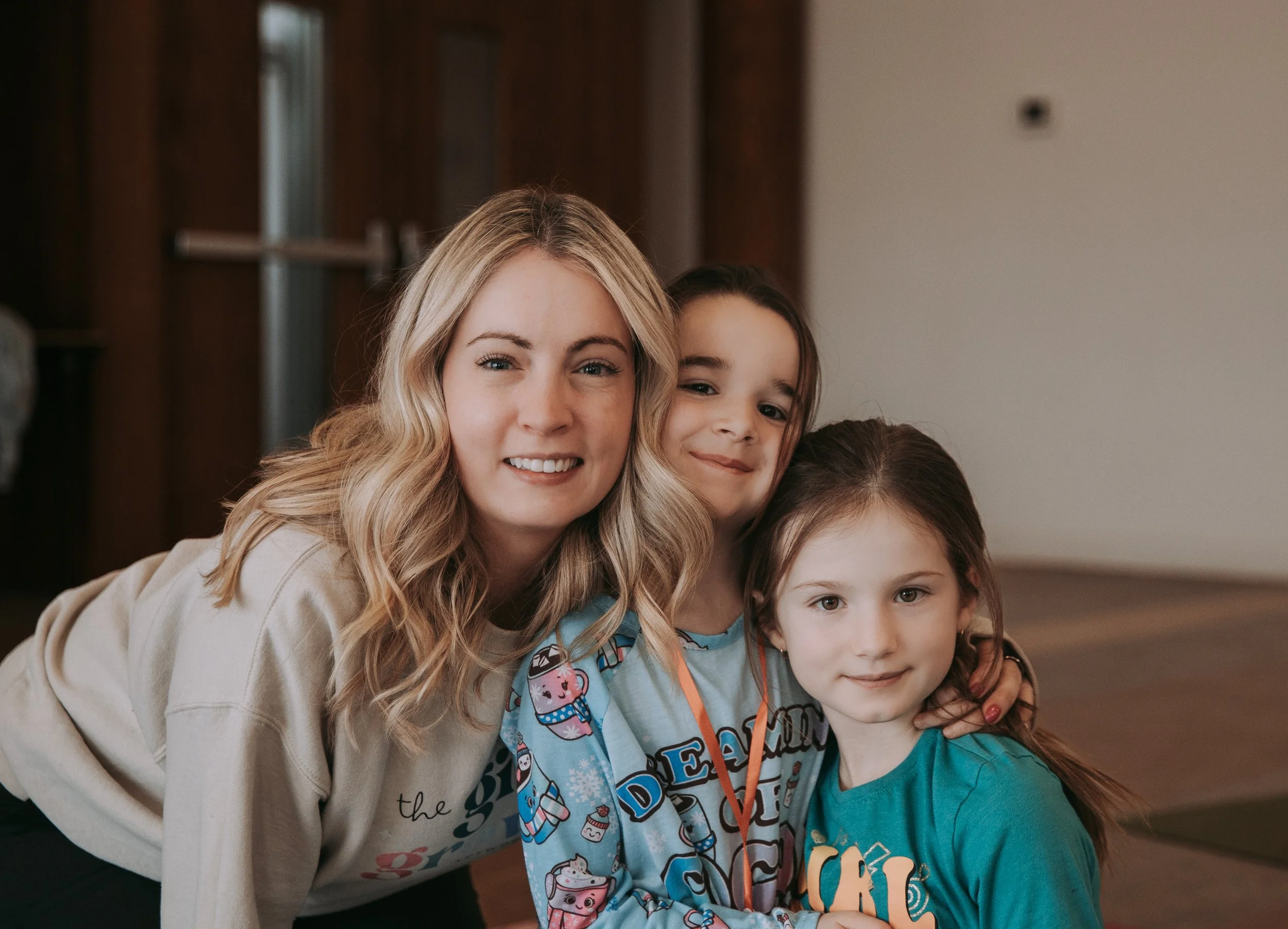 A woman with long blonde hair and three young girls, all smiling and posing together indoors.