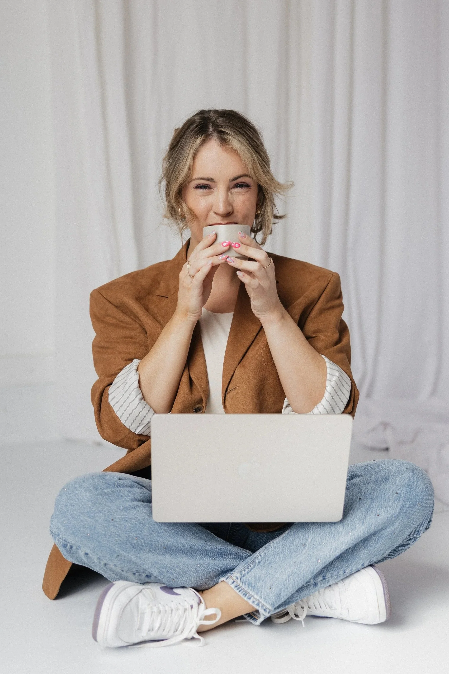 A woman sitting cross-legged on the floor, wearing a brown blazer, light-colored shirt, and blue jeans, holding a white mug and looking at a laptop in front of her.