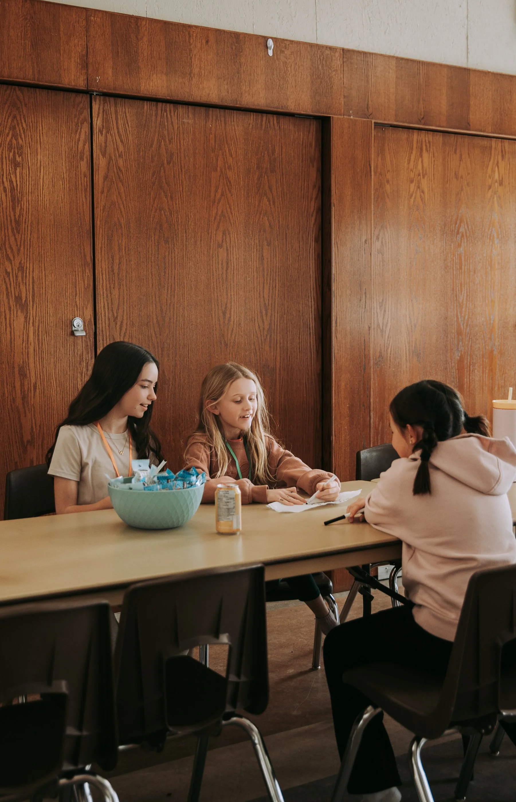 Four girls sitting at a table engaging in conversation. The table has snacks and drinks, with a wood-paneled wall in the background.