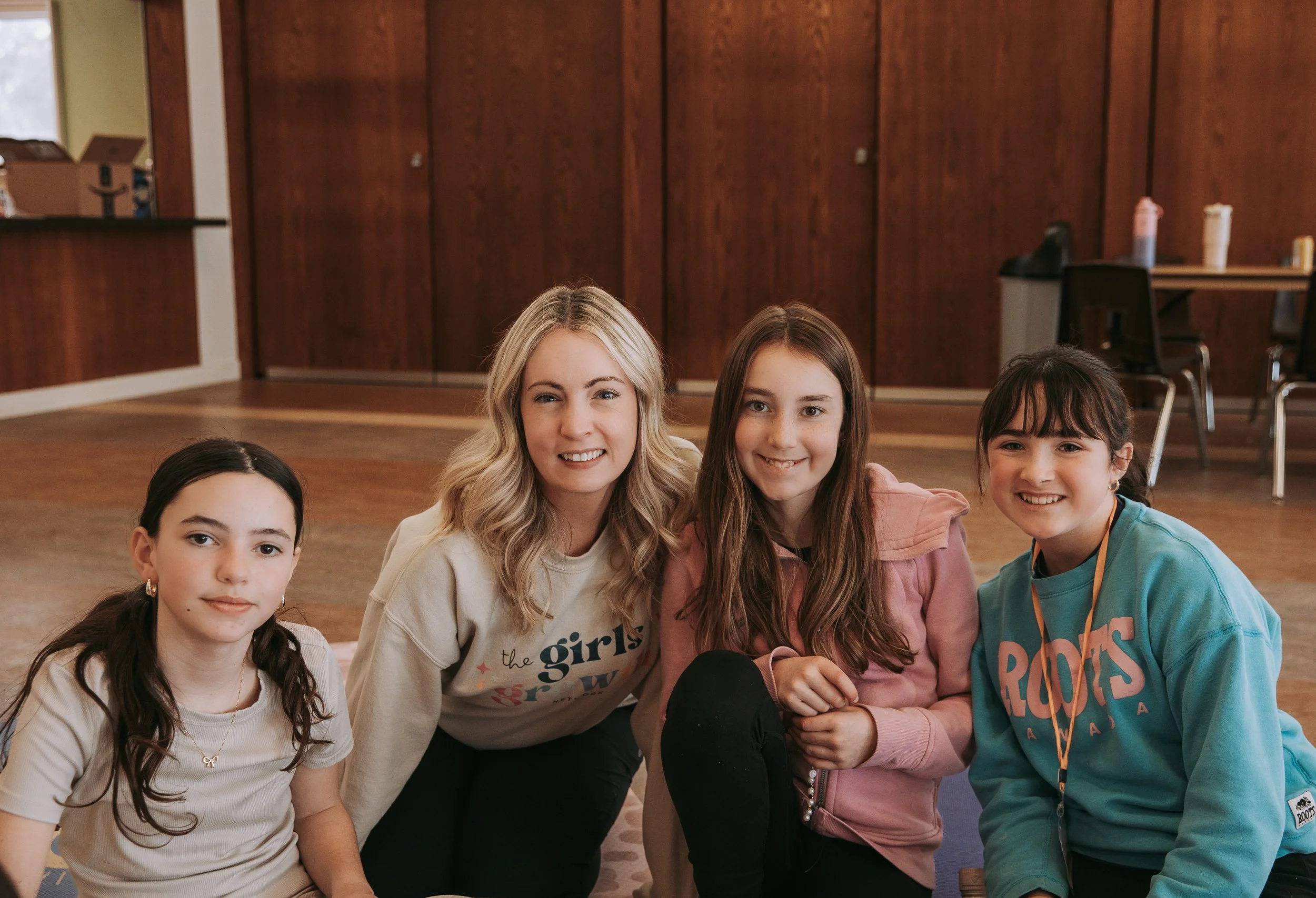Four girls and one woman pose for a photo in a room with wooden walls and a table in the background. They are smiling and sitting close together on the floor.