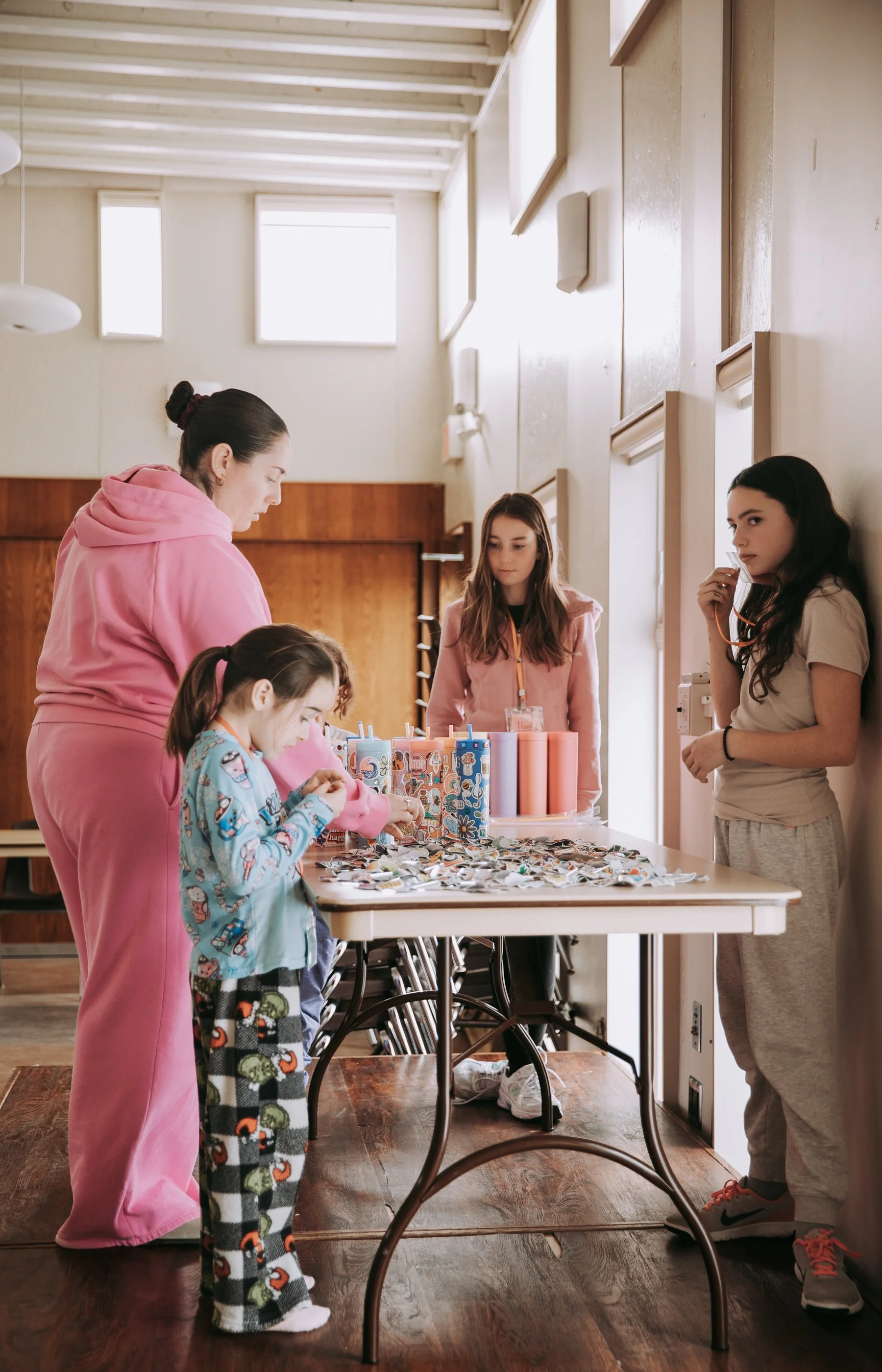 Adult woman in pink hoodie and pajamas, young girl in colorful pajamas, and teenage girl in casual clothes, standing around a table with various craft supplies in a bright room.