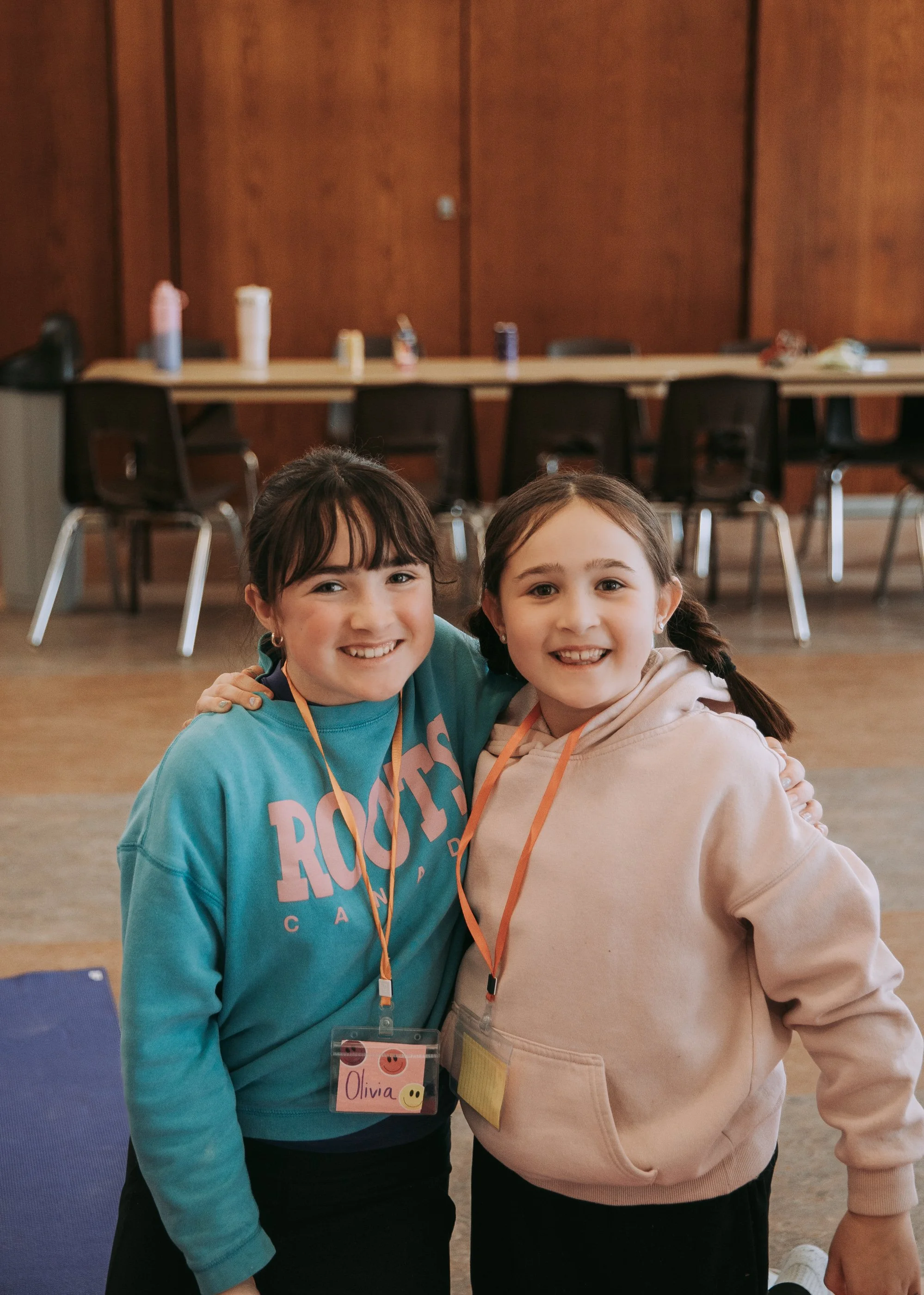 Two smiling young girls standing close together indoors, with a wooden wall and chairs in the background. They are wearing casual clothing and name tags, one with the name Olivia.