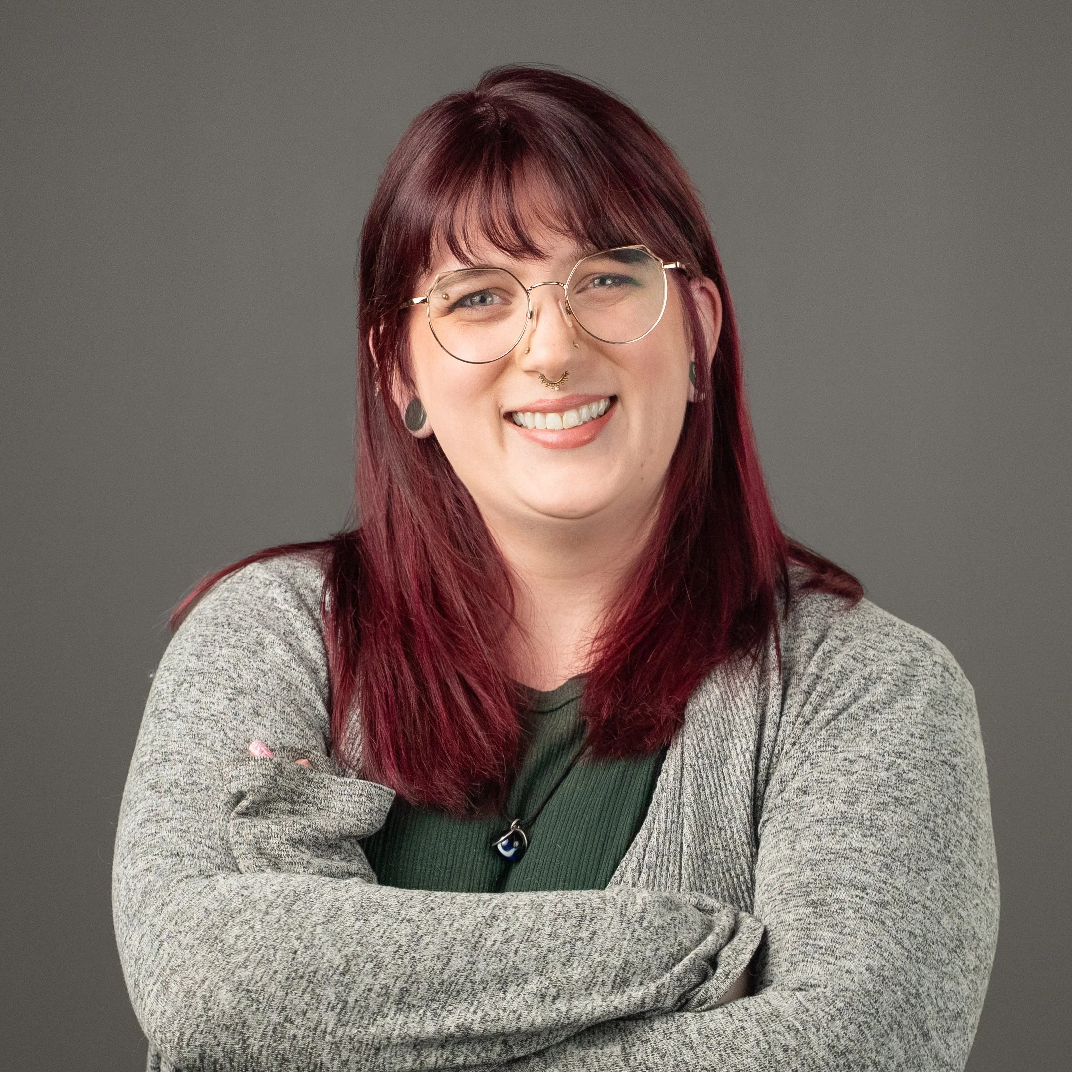 Close-up of a woman with red hair, wearing glasses, smiling, with a gray background, wearing a gray sweater over a green shirt.