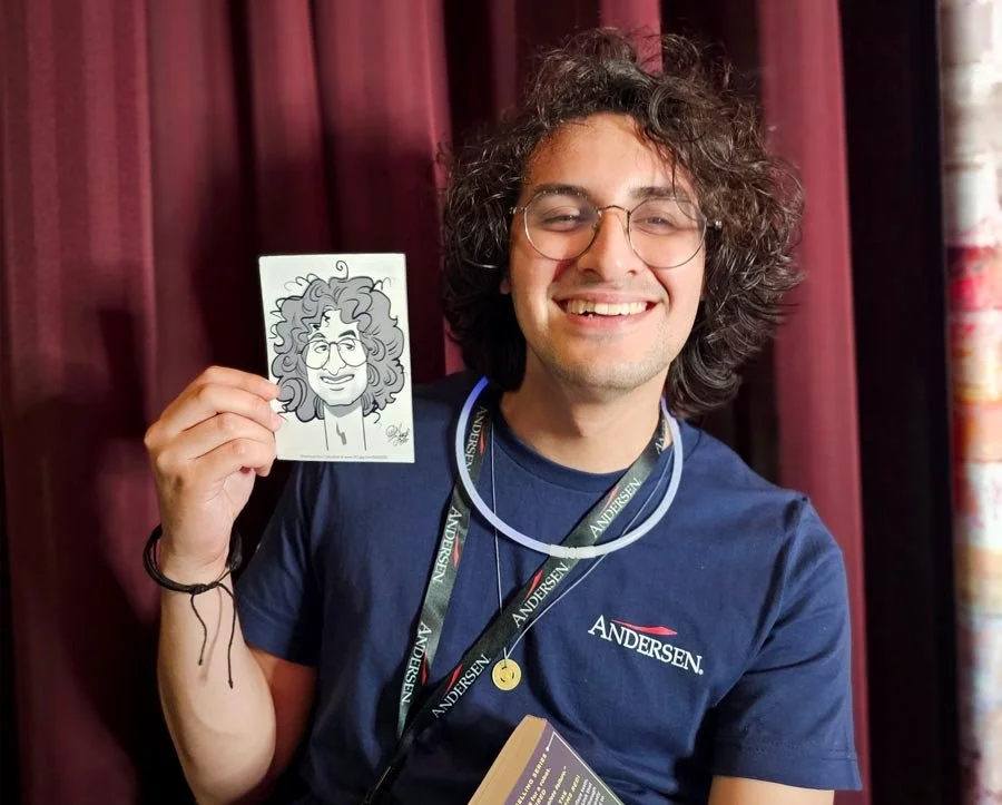 A young man with curly hair and glasses smiling while holding a caricature portrait of himself. He is wearing a navy blue Andersen shirt, a lanyard, and a ring with a yellow smiley face. He is standing in front of a maroon curtain.