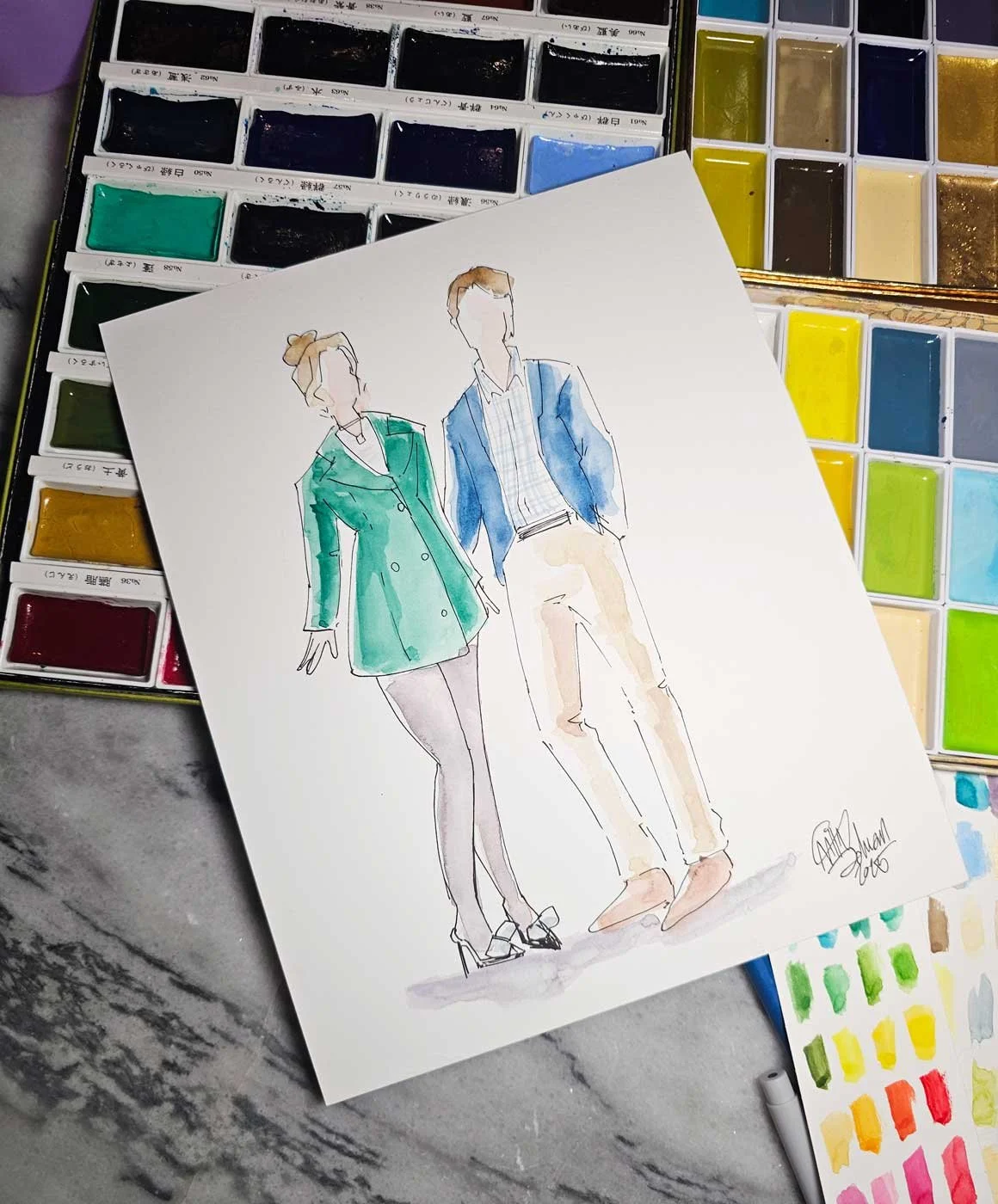 A painting of a woman with a green coat dress and a man with a blue blazer & khaki slacks rests on a watercolor palette and marble table.