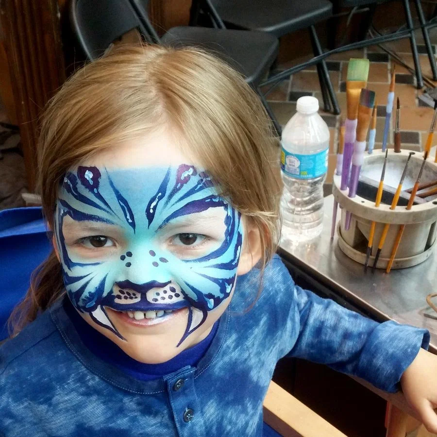 A girl is smiling and wearing a blue tiger face paint.