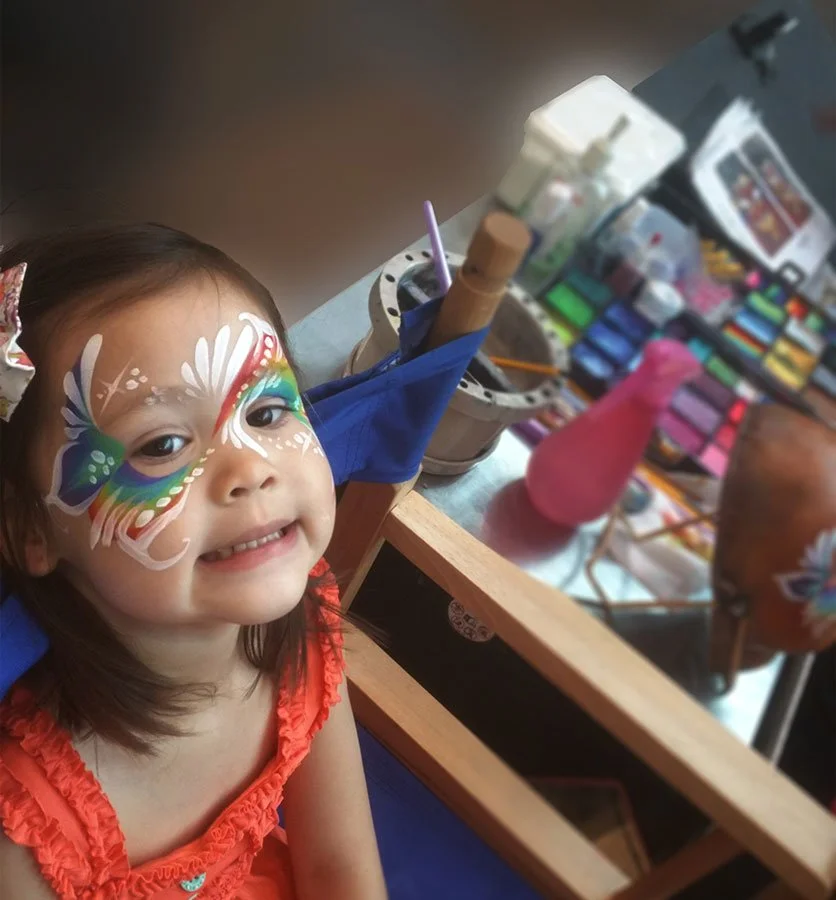 A girl is wearing rainbow face paint with paints in the background.