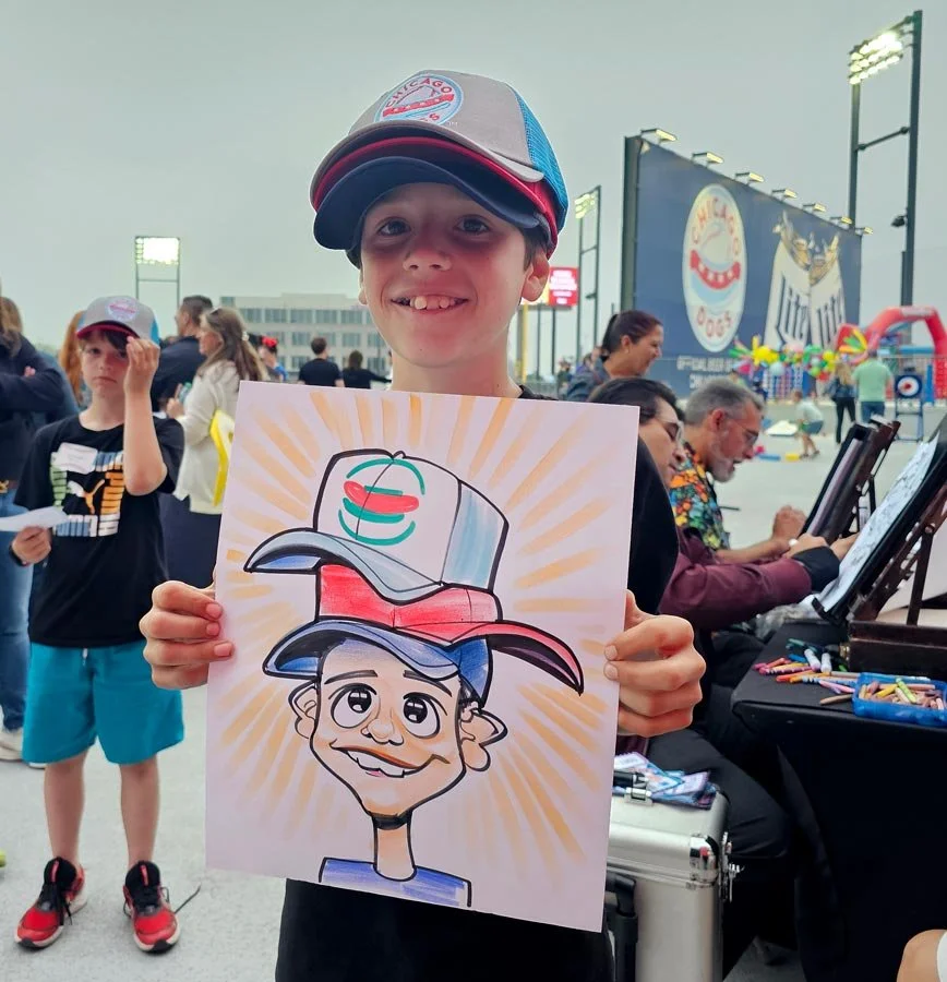 A smiling boy wearing 3 baseball caps holds up his caricature of him with exaggerated hats.