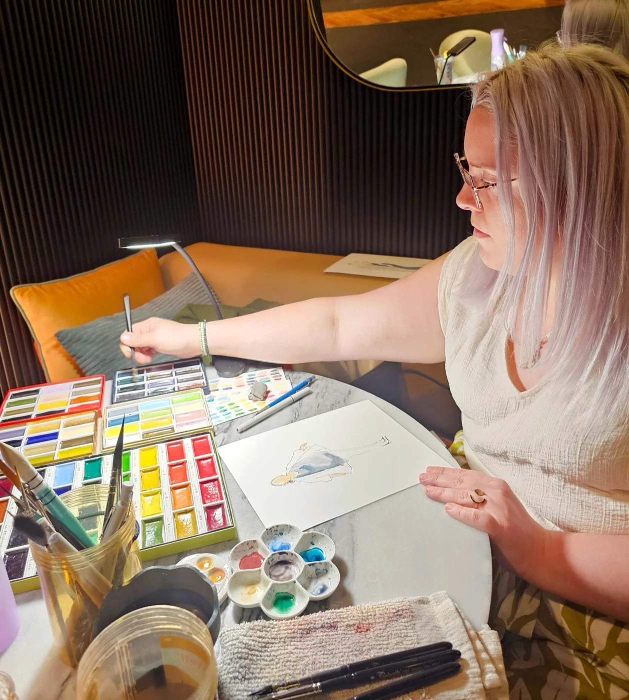 An artist is sitting at a table with an assortment of watercolors and a desk lamp and is painting a woman.