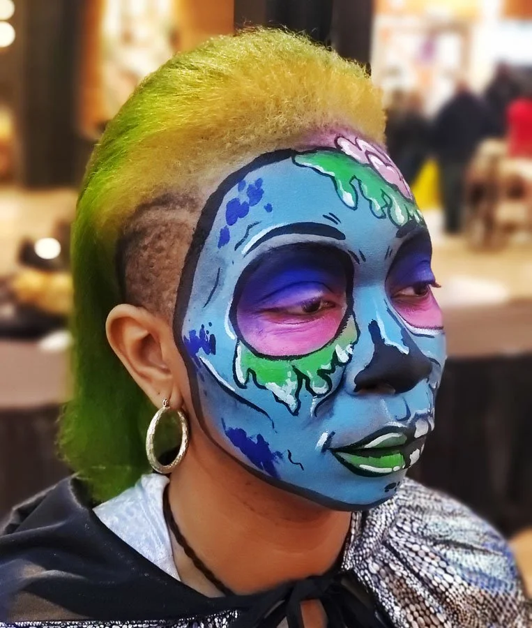 A woman with yellow and green hair wears a cartoon-ish zombie face paint.