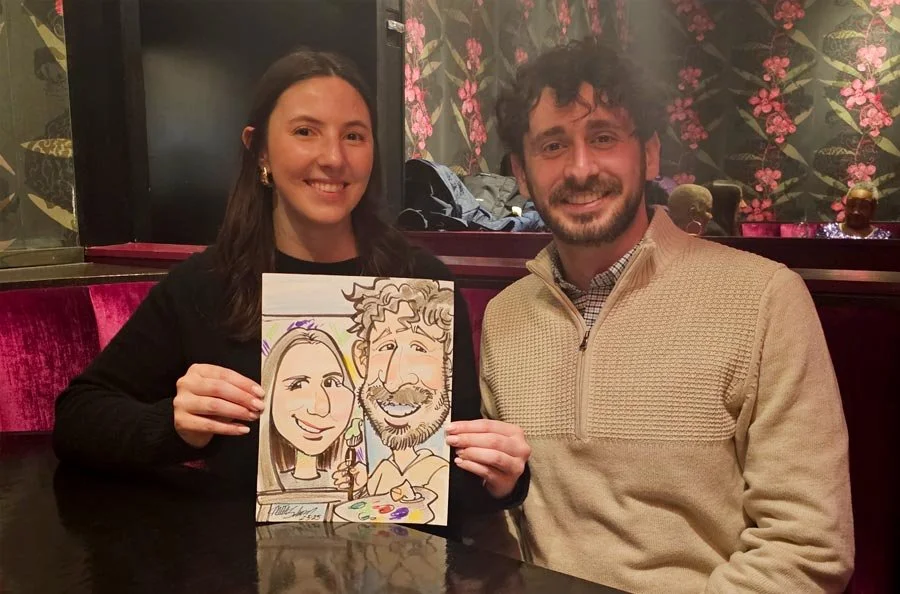 A smiling couple holds their caricature of the man painting the woman.