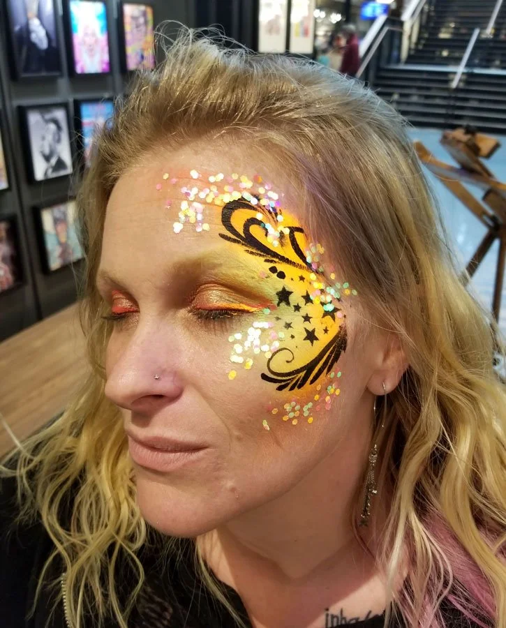 A woman is wearing a yellow face paint with black stars and swirls.