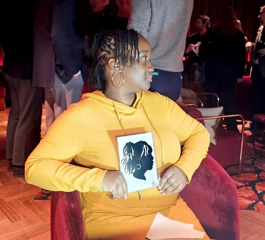 A woman wearing a yellow dress sits in a red velvet chair and holds her silhouette.