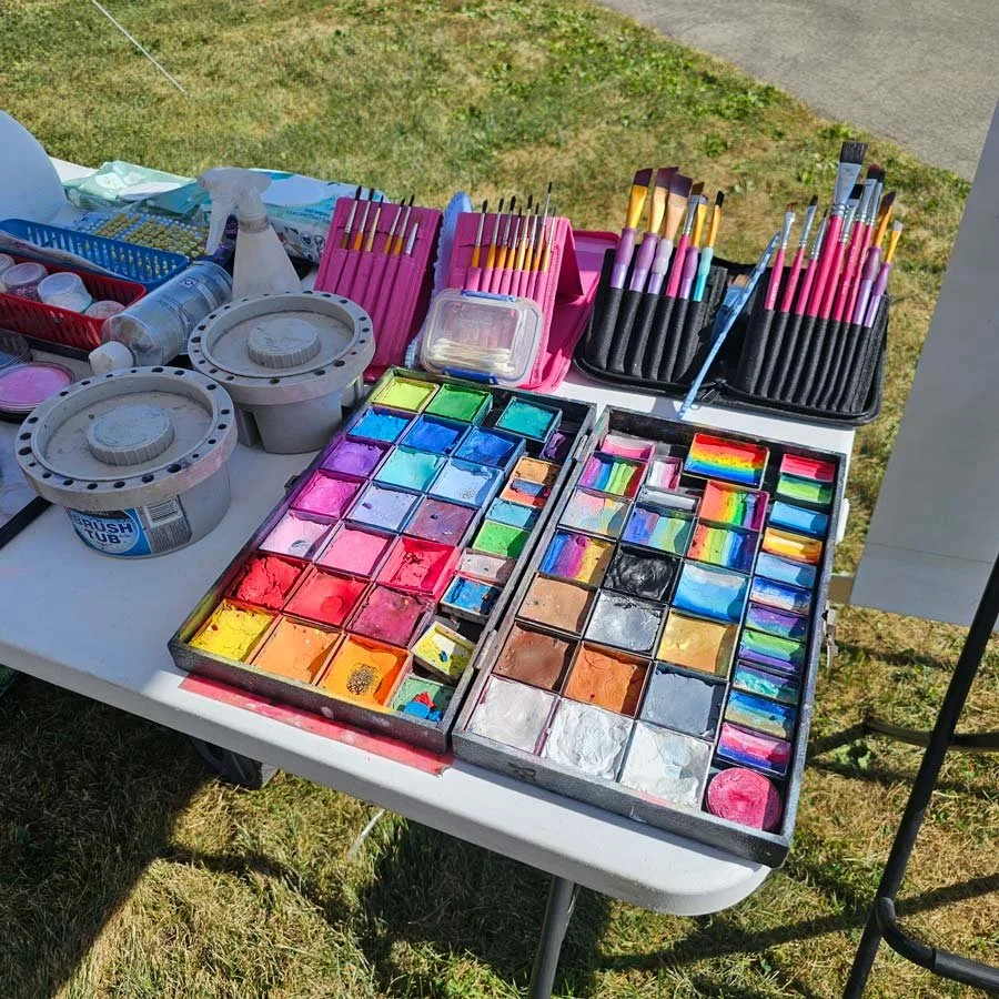 A case of bright colorful face paints are on a table next to a collection of brushes and water containers.