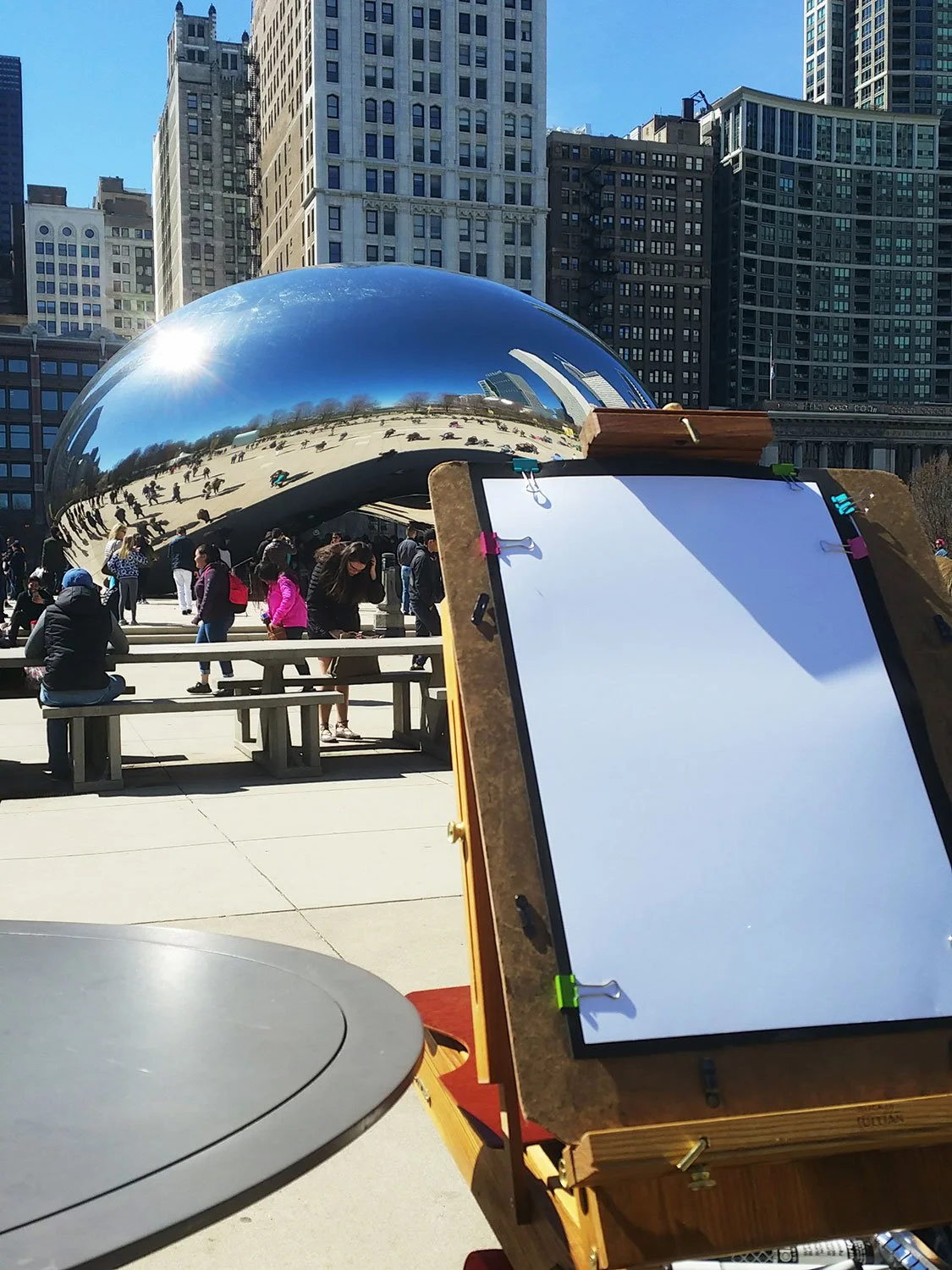 People gathered at Millennium Park in front of the Cloud Gate sculpture in Chicago on a sunny day, with an artist setup in the foreground.